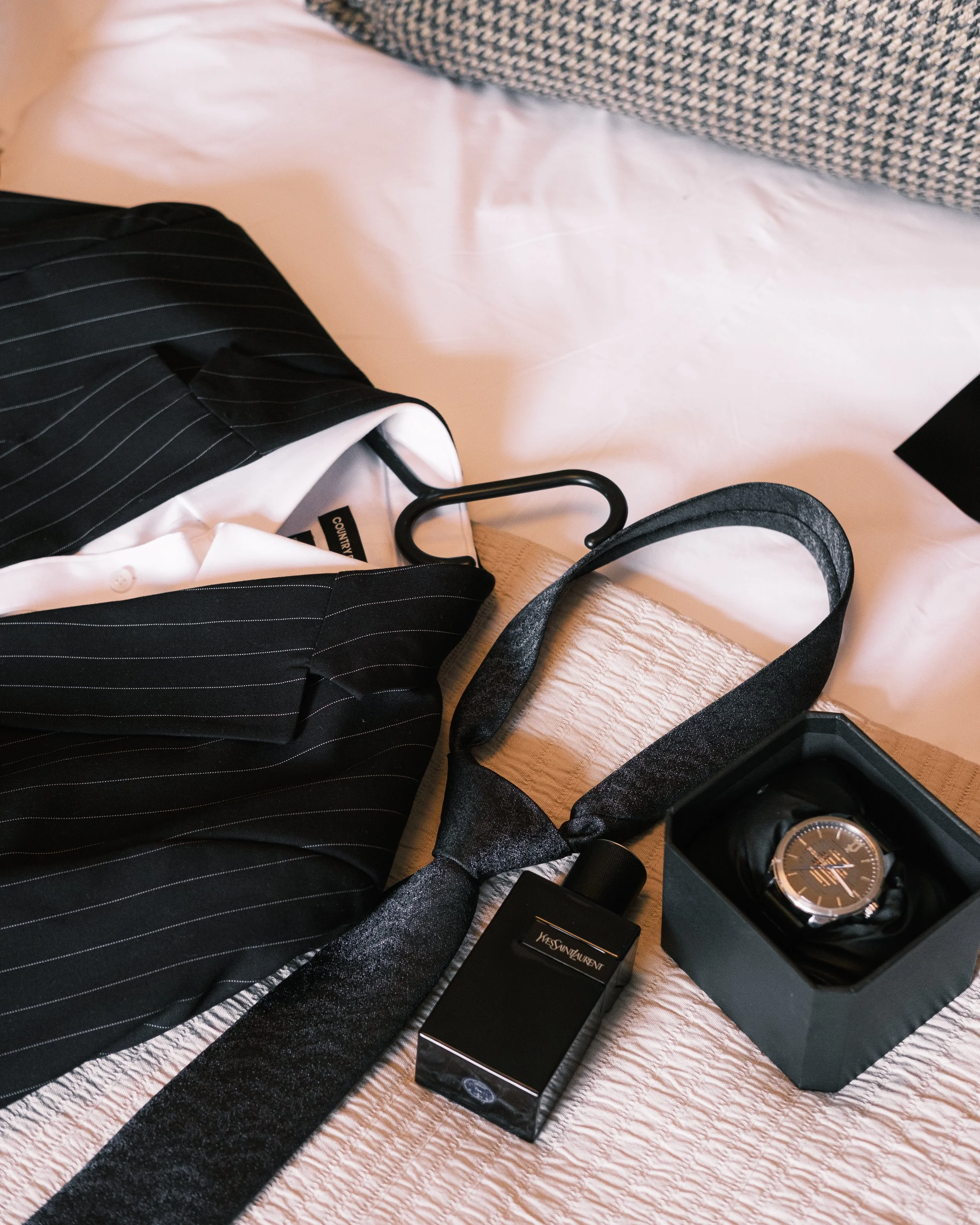 Taylor of a black wallet, a black bottle of Yves Saint Laurent perfume, a black wristwatch in a black box, a black and silver necktie, and a pinstripe blazer on a white bedspread.