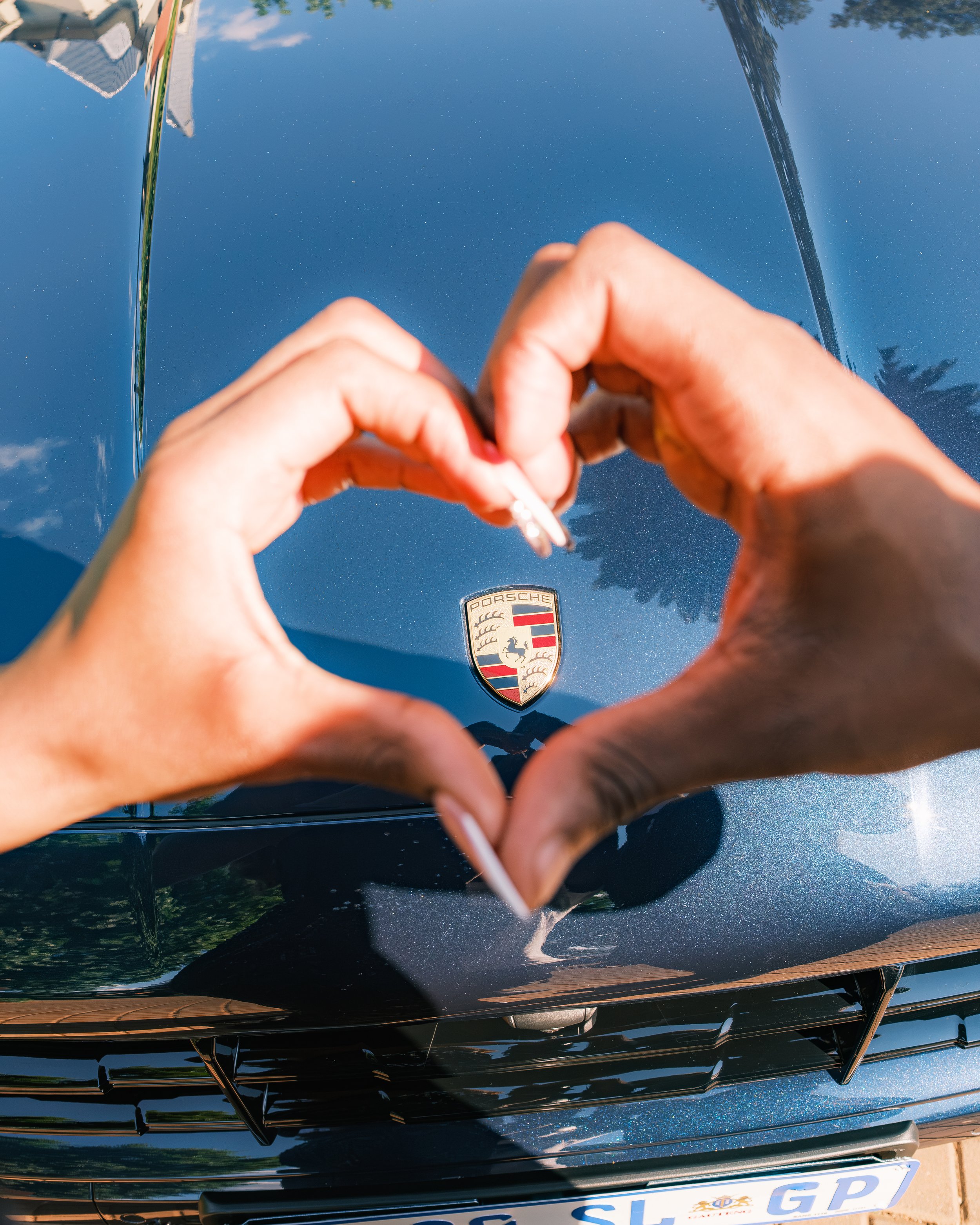 Person making a heart shape with their hands around the Porsche logo on the hood of a black car, which reflects the blue sky and trees.