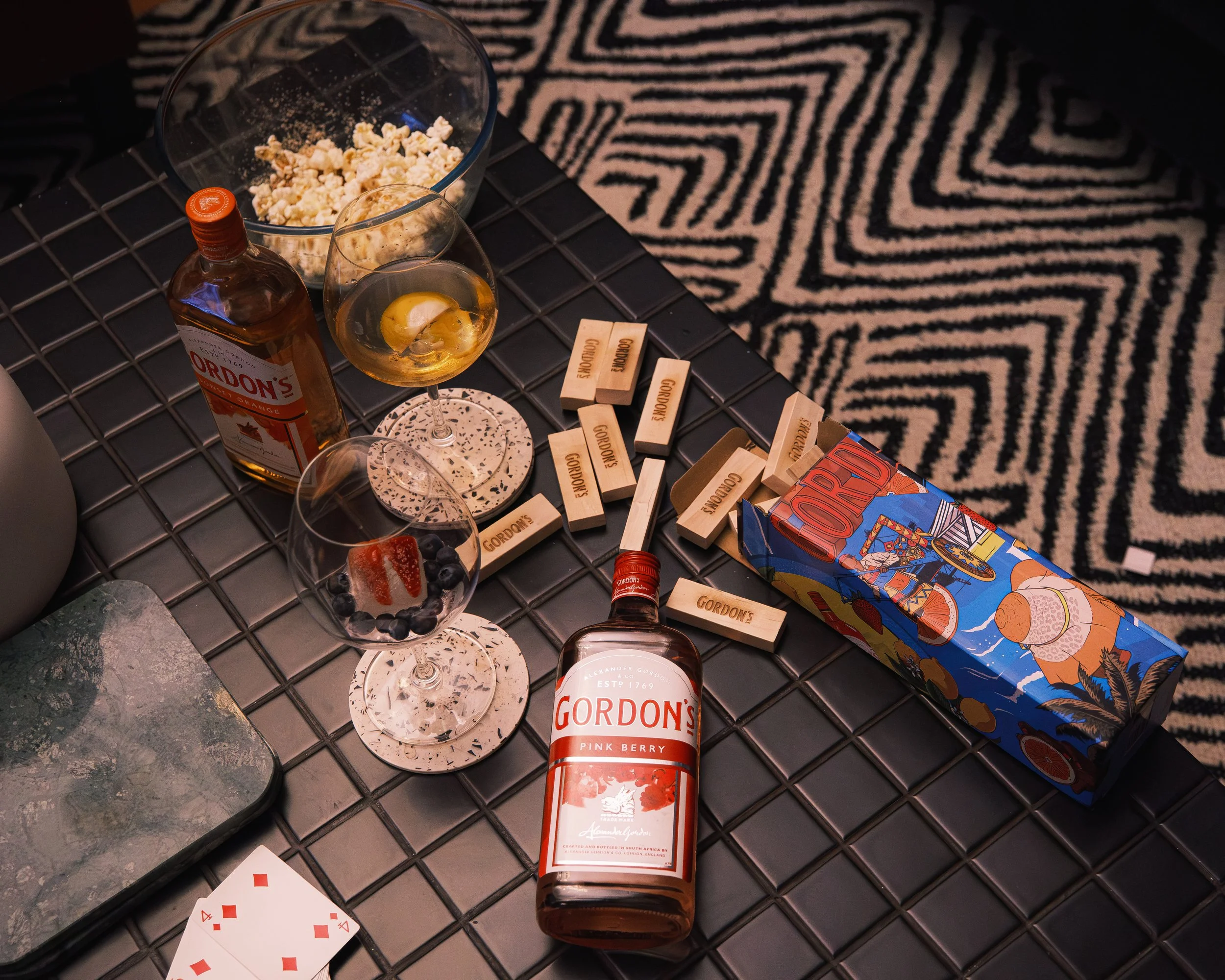 A black tiled table with a bottle of Gordon's Pink Berry gin, two glasses with drinks, a box of Gordon's tonic, a bowl of popcorn, a glass with blueberries and a strawberry, a deck of playing cards, wooden dominoes, and coasters, with a black and whi
