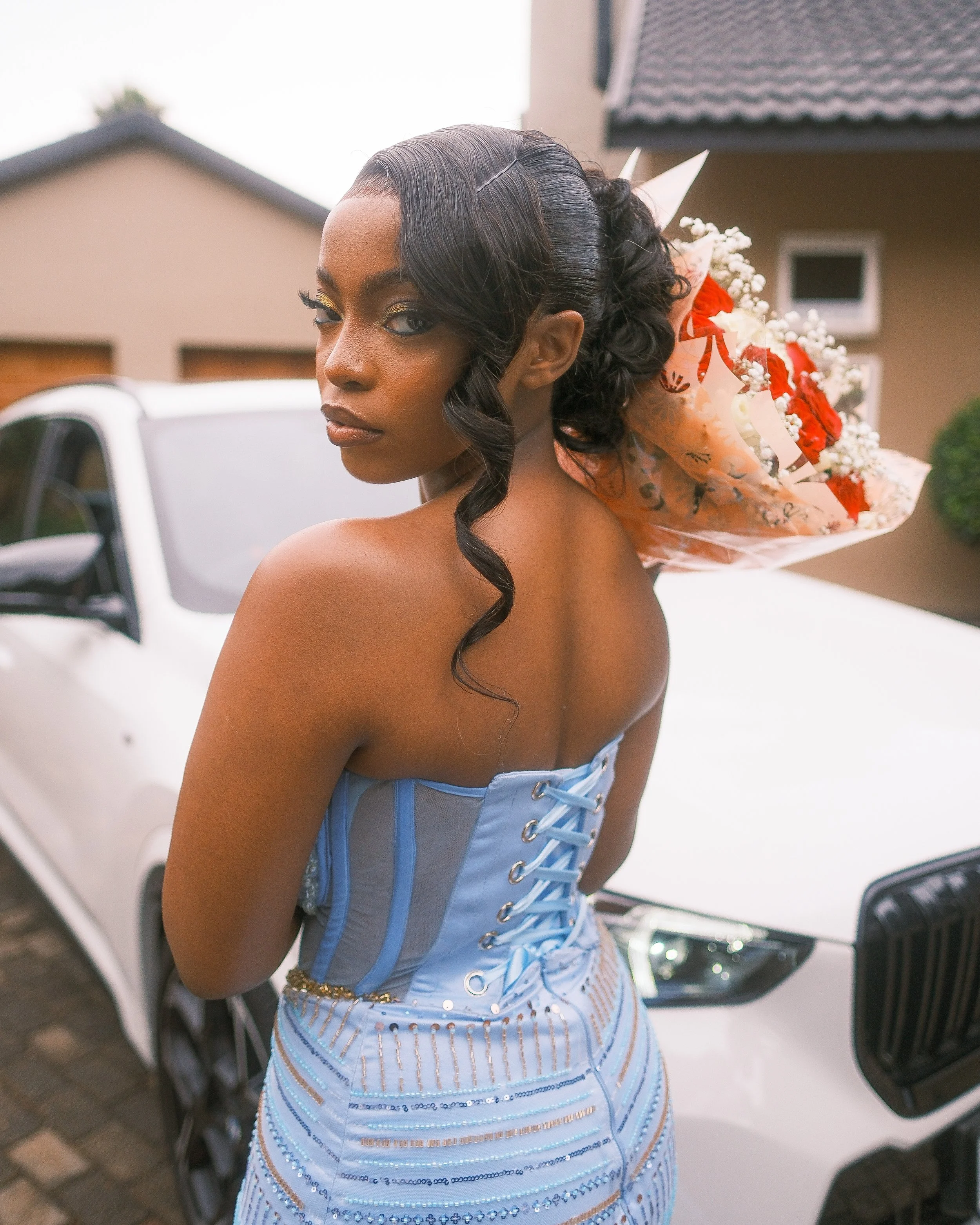 A woman with dark skin, styled hair, and makeup, wearing a strapless light blue corset dress, standing outdoors near a white car, holding a bouquet of red and white flowers.
