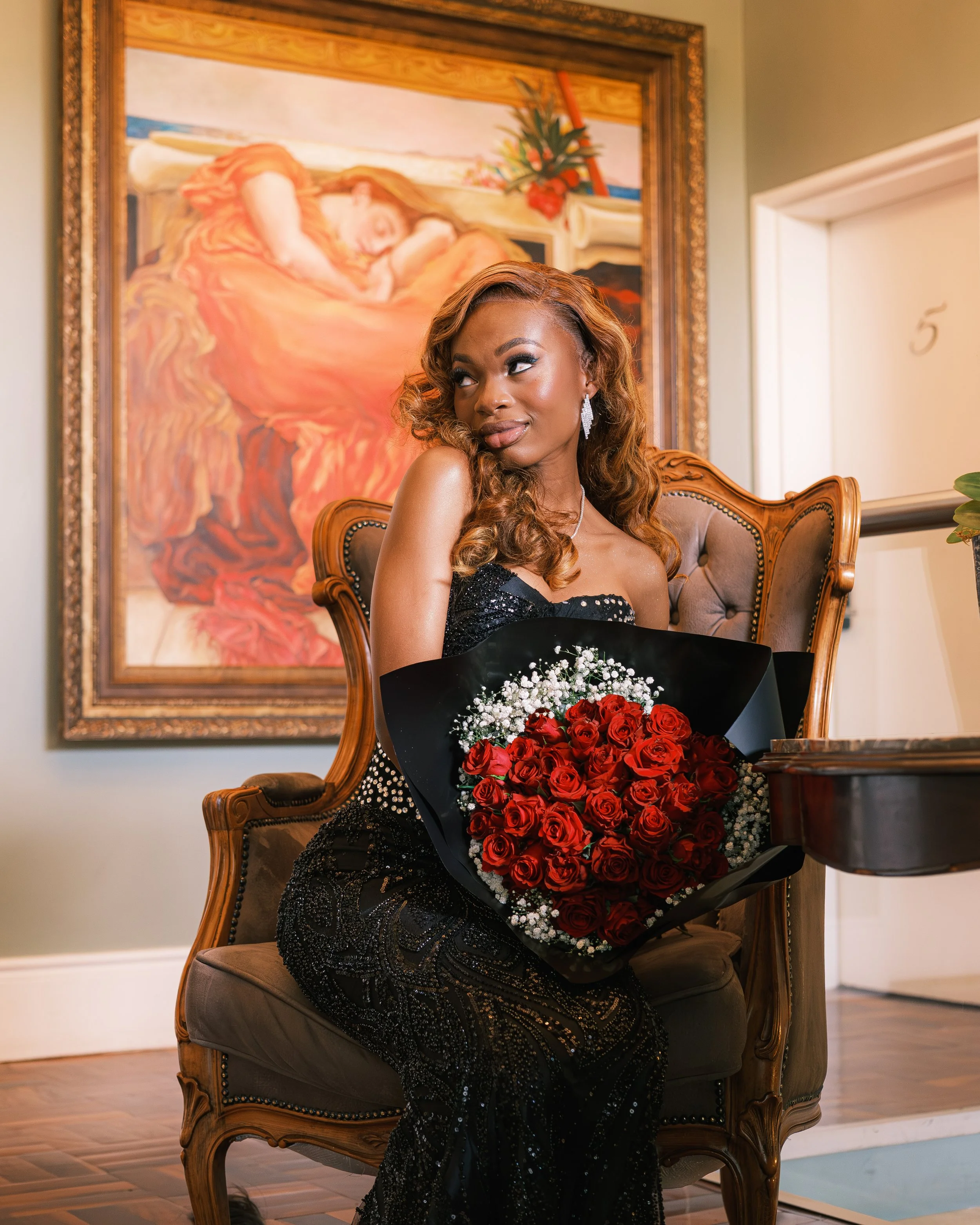 A woman with long, curly hair dressed in a black, beaded gown sitting on a vintage armchair, holding a bouquet of red roses, in an elegant room with a painting of a woman sleeping on the wall behind her.