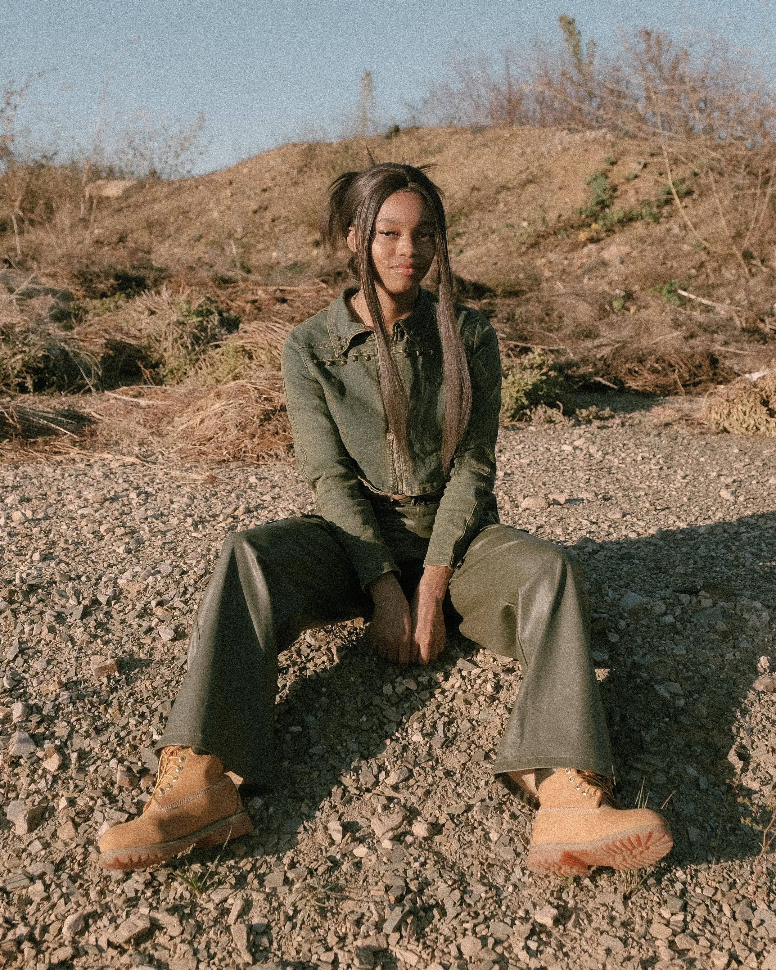 A woman with long dark hair, wearing a green jacket, green wide-leg pants, yellow boots, and sitting on gravel ground in an outdoor area with a dirt hill and sparse bushes in the background.