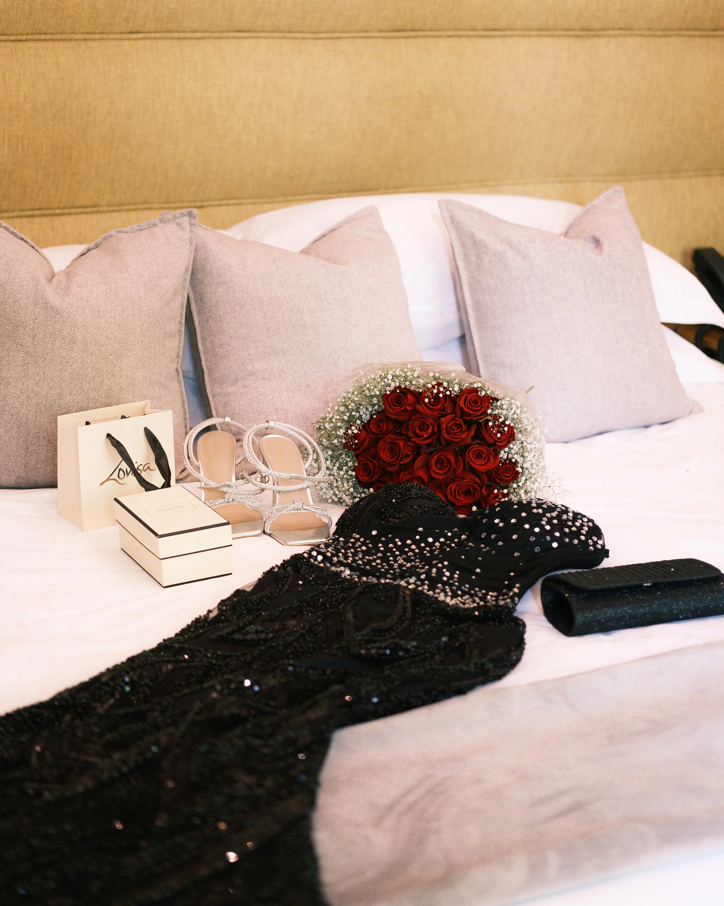 A hotel bed with a bouquet of red roses, a pair of silver high heels, a black dress with sequins, black clutch purse, and jewelry on it.