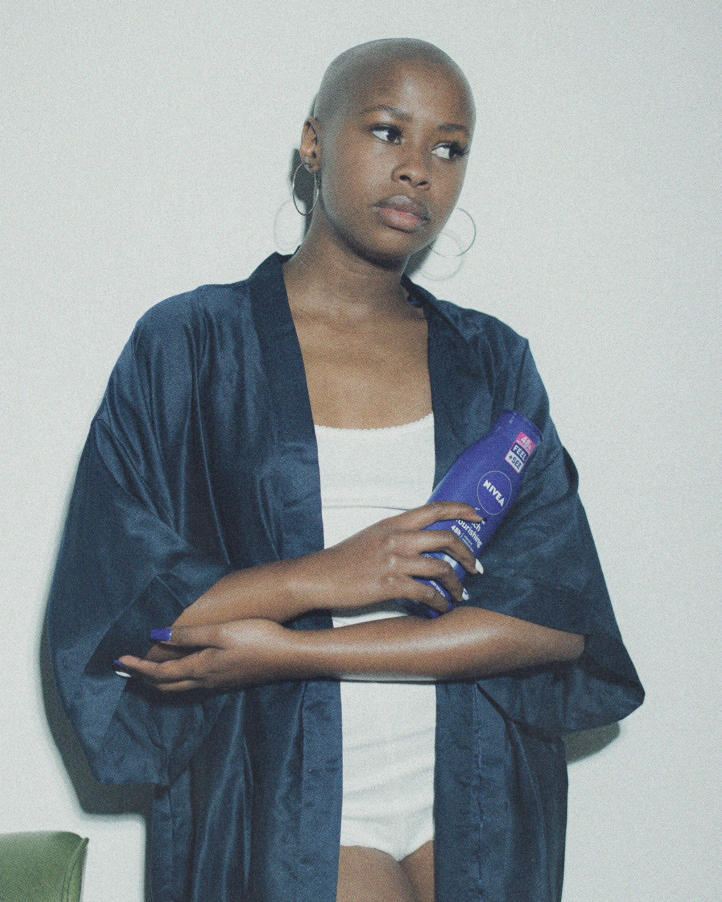 A woman with a bald head, wearing hoop earrings and a navy robe over a white top, holding a bottle of Nivea lotion, standing against a plain wall.