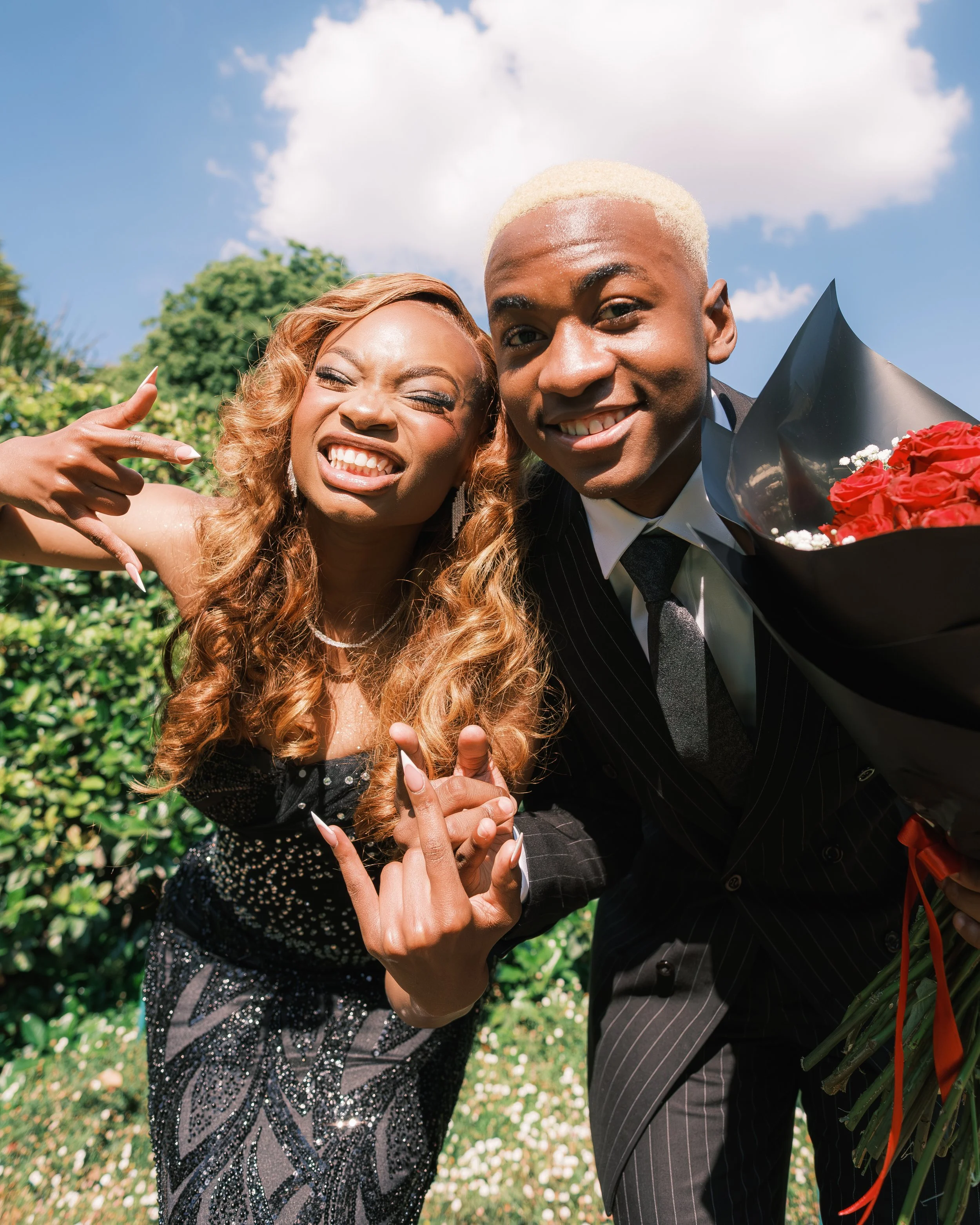 A young man in a black pinstripe suit holding a bouquet of red flowers, smiling next to a young woman with long curly hair making a rock sign with her hand, both enjoying a sunny outdoor day.