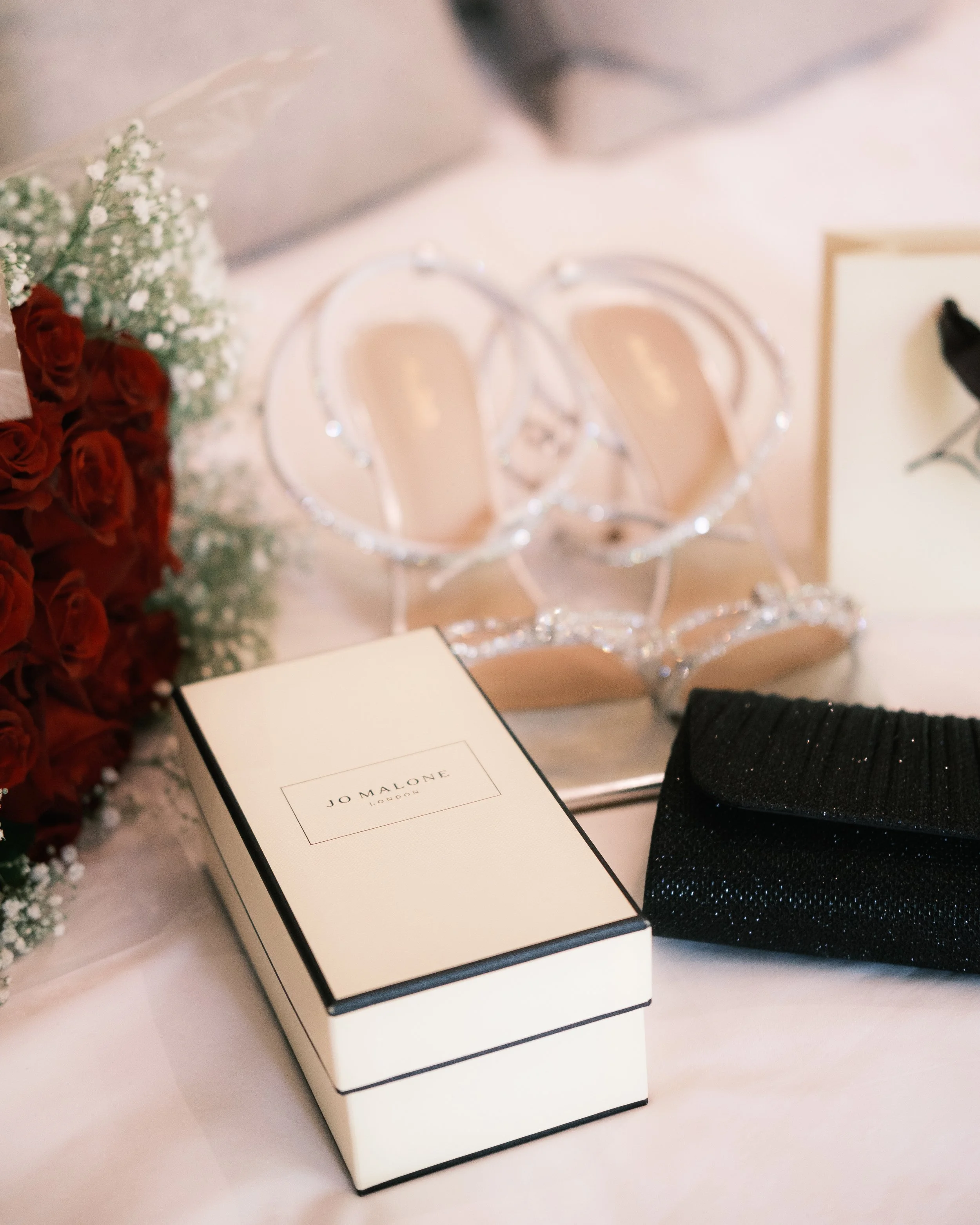 A pair of embellished high-heeled shoes with rhinestones, a black clutch, a white box from Jo Malone London, some red roses and white baby's breath flowers, and a blank card with a black flower design.