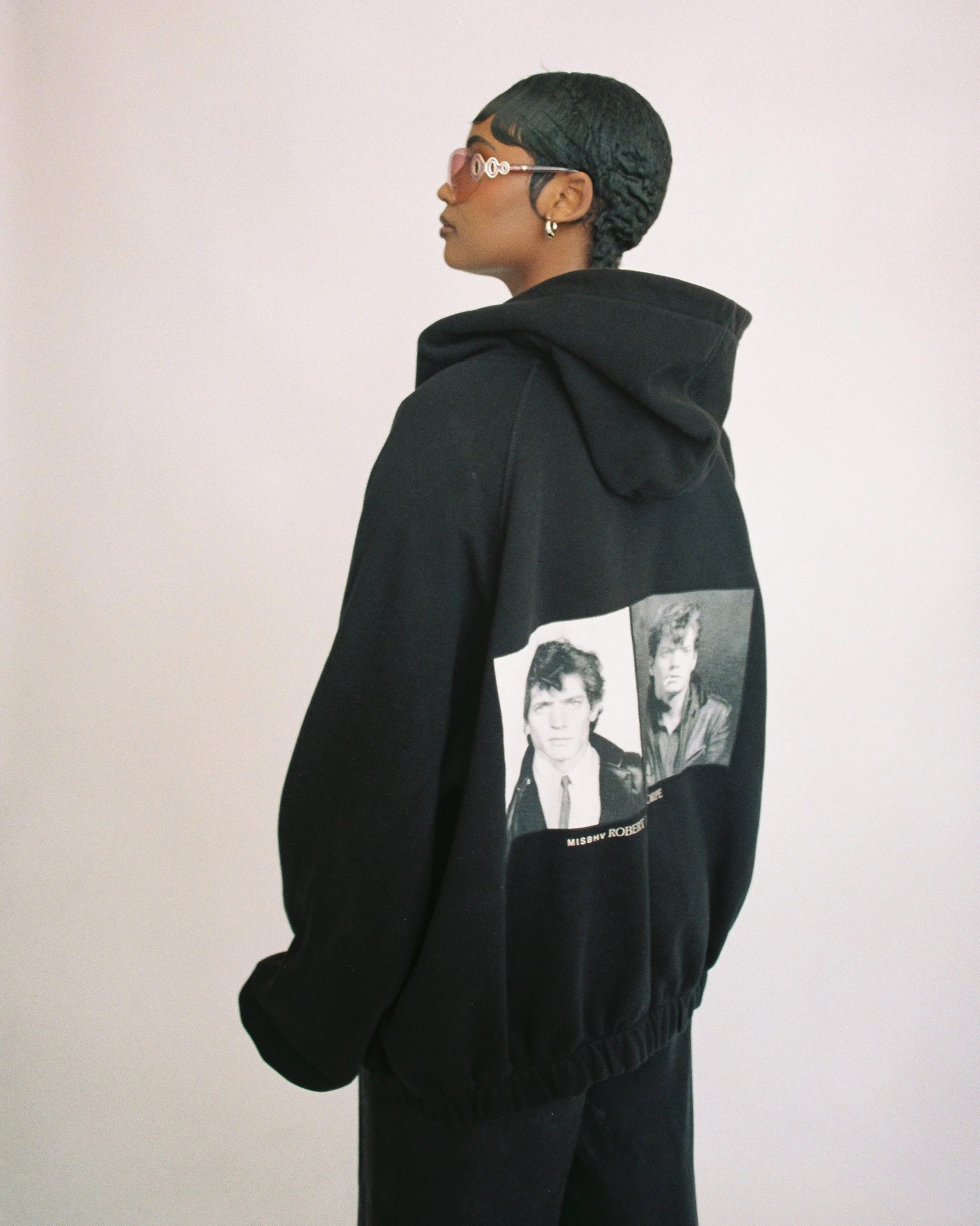Person wearing a black hoodie with a graphic print of two men on the back, sunglasses, and earrings, standing against a plain background.