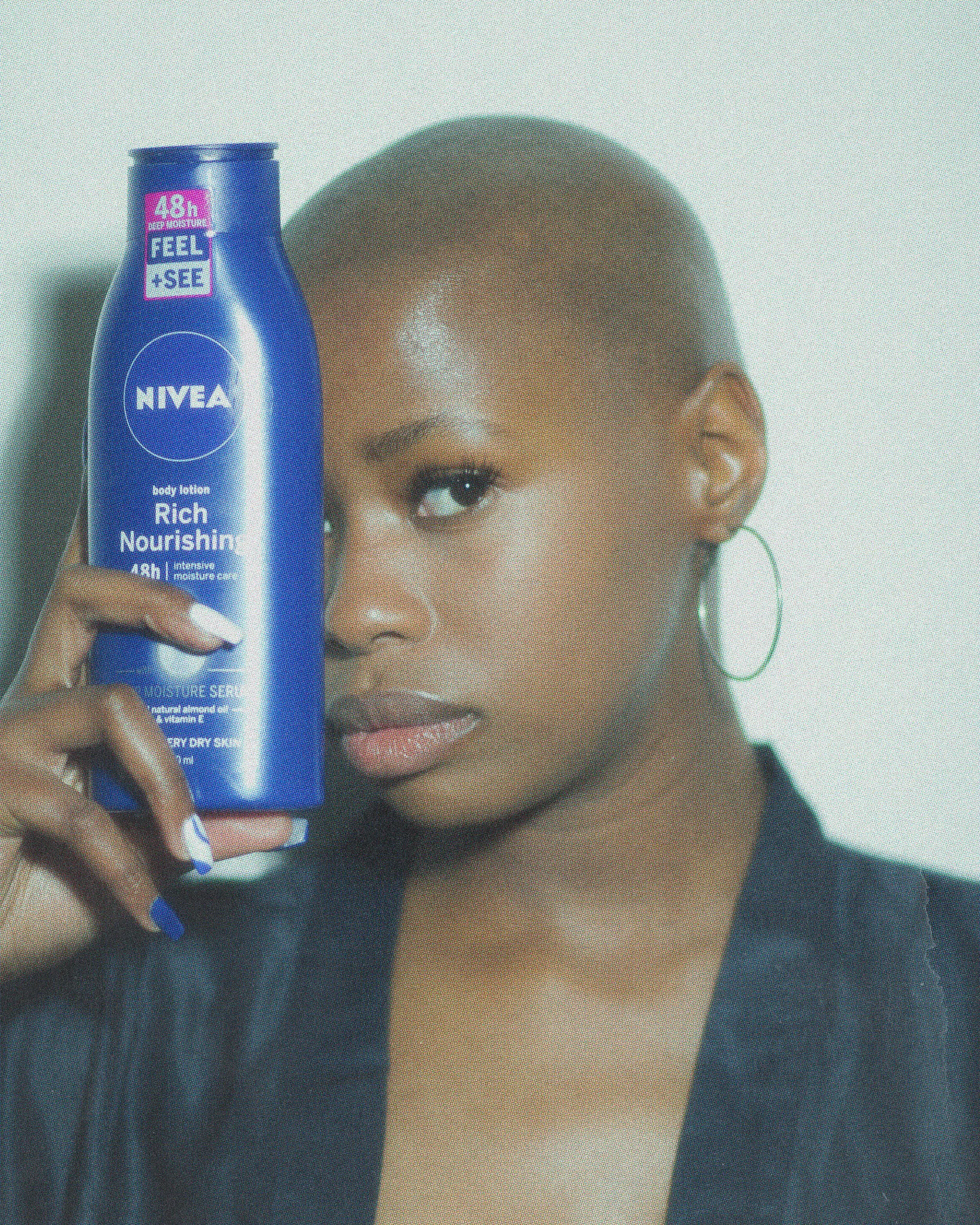 A woman holding a blue Nivea body lotion bottle in front of her face, partially covering her right eye, with a neutral expression and hoop earrings.
