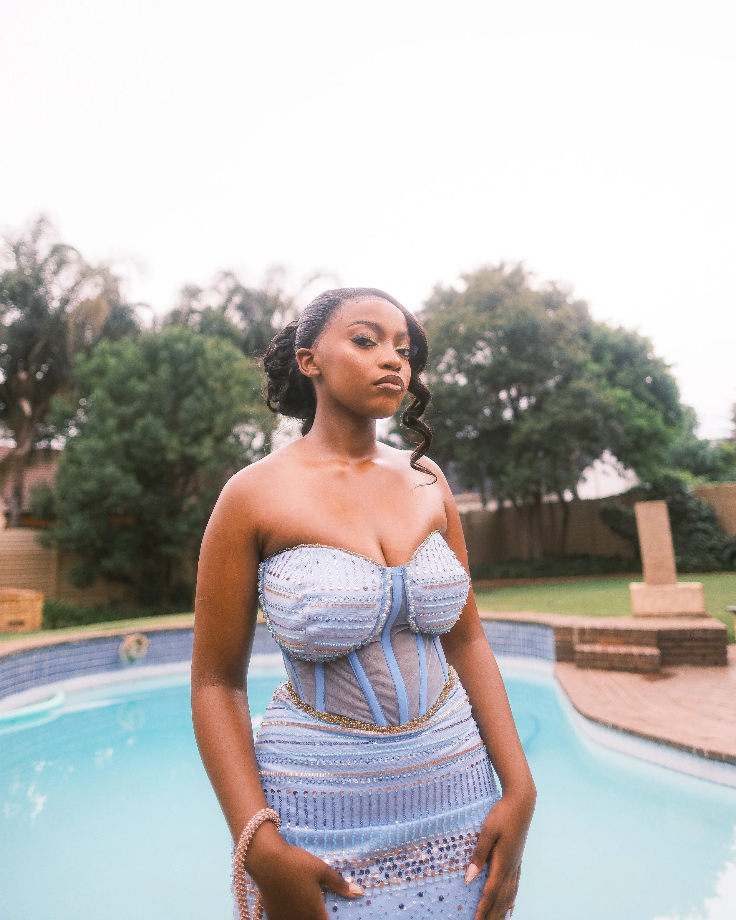A woman in a strapless blue dress with beads and sequins, standing by a backyard pool with trees and fence in the background.