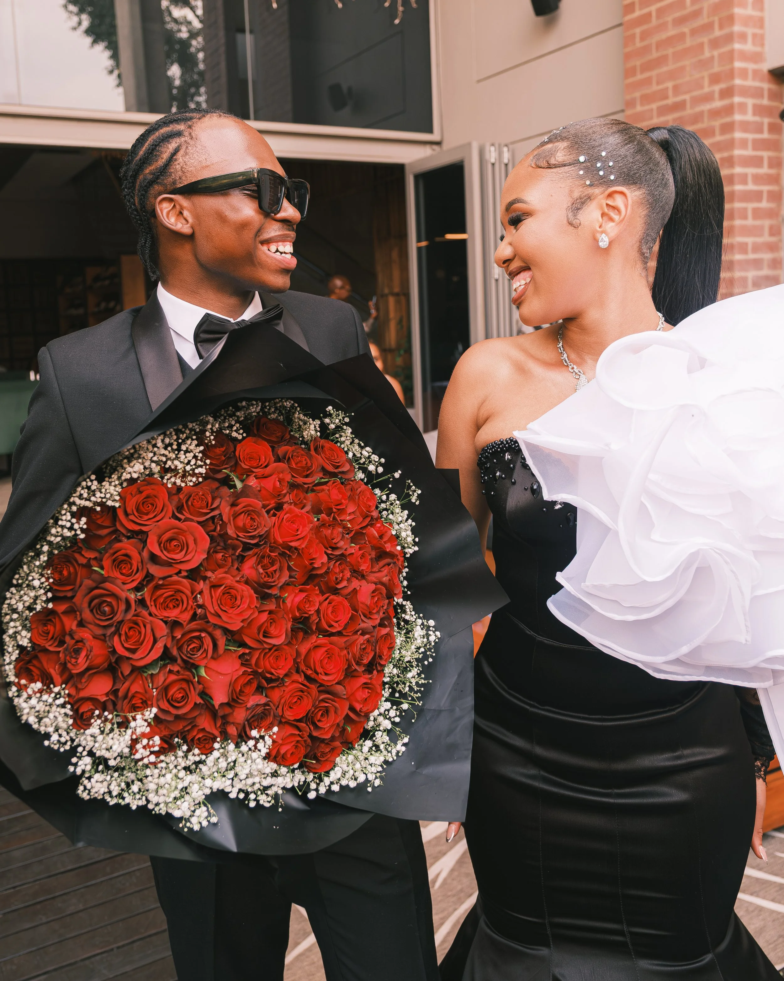 A man in a tuxedo with sunglasses holding a large bouquet of red roses, smiling at a woman in a black strapless dress with jewelry, at a building entrance.