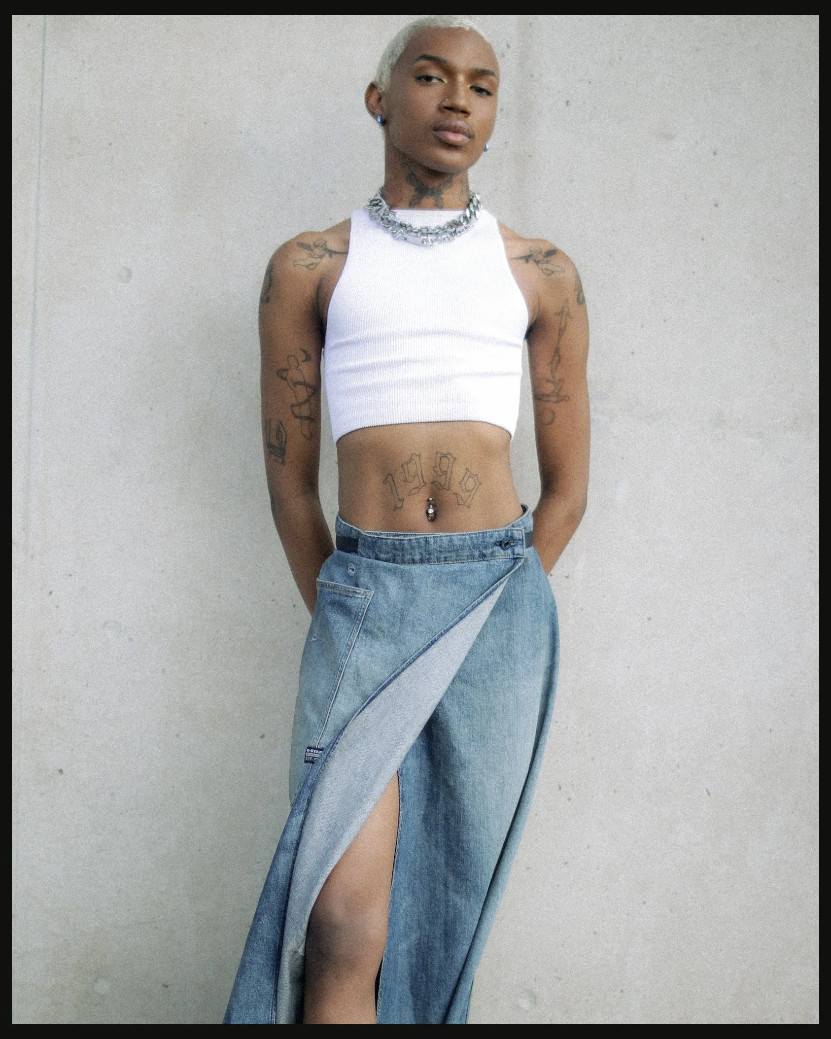 A young woman with platinum blonde hair standing against a plain wall, wearing a white crop top, wide-legged jeans with a high slit, accessorized with necklaces, earrings, tattoos on arms and stomach, and a belly button piercing.