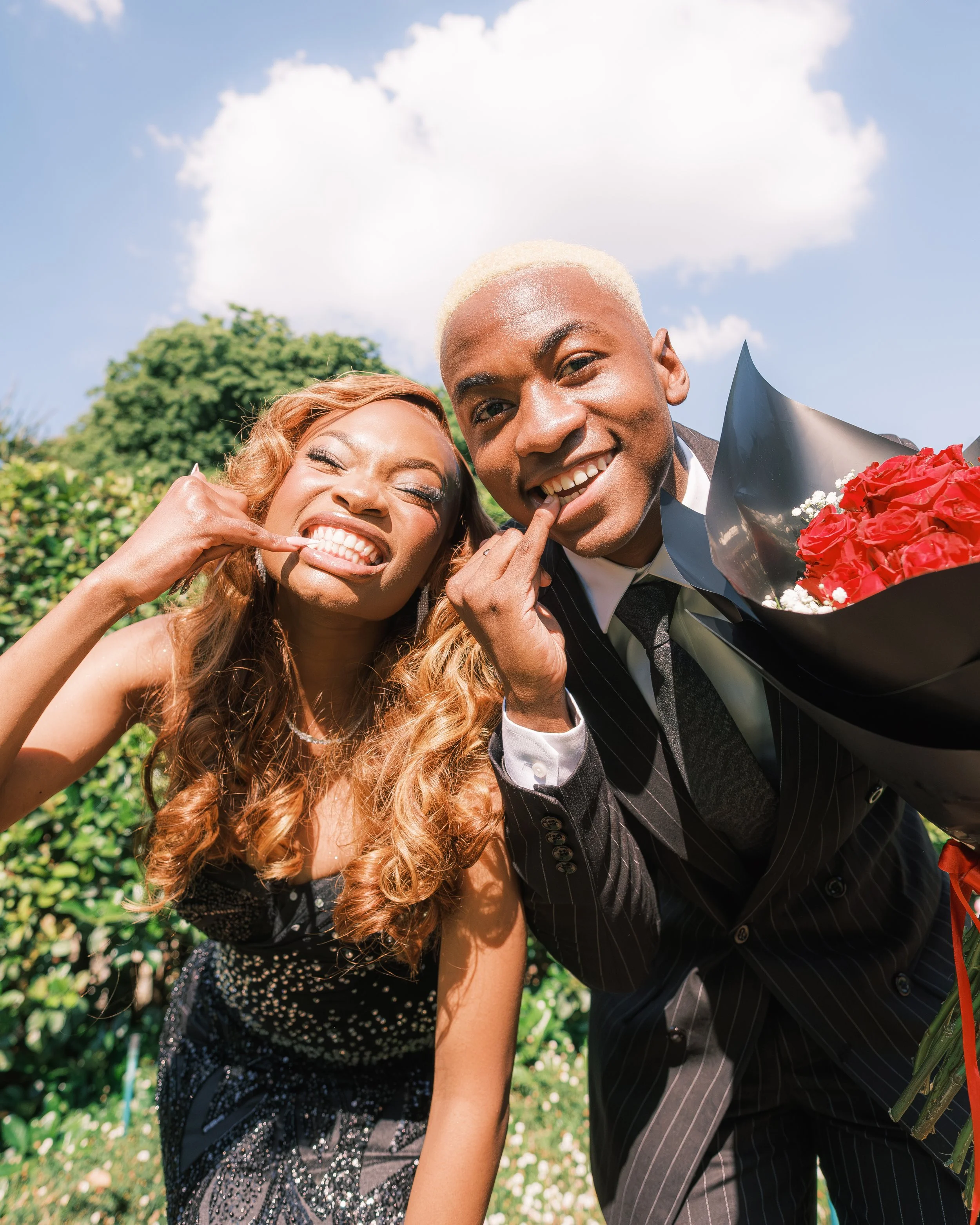 A happy couple in formal wear making funny faces outdoors with blue sky and greenery in the background, the woman with long curly hair and the man holding a bouquet of red roses.