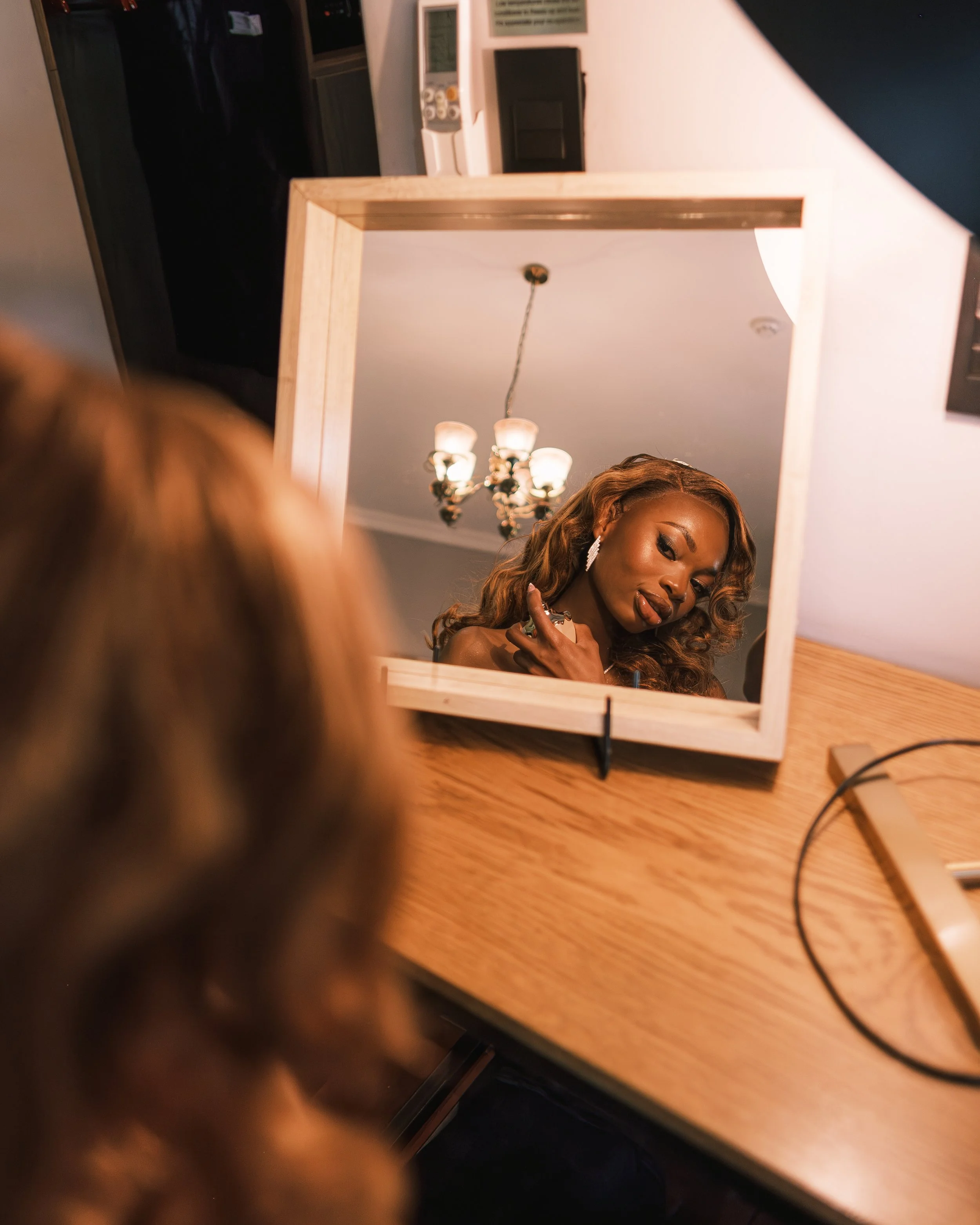 A woman looking into a square mirror on a wooden table, with a chandelier visible in the reflection, and a few electronic devices on the countertop in the background.
