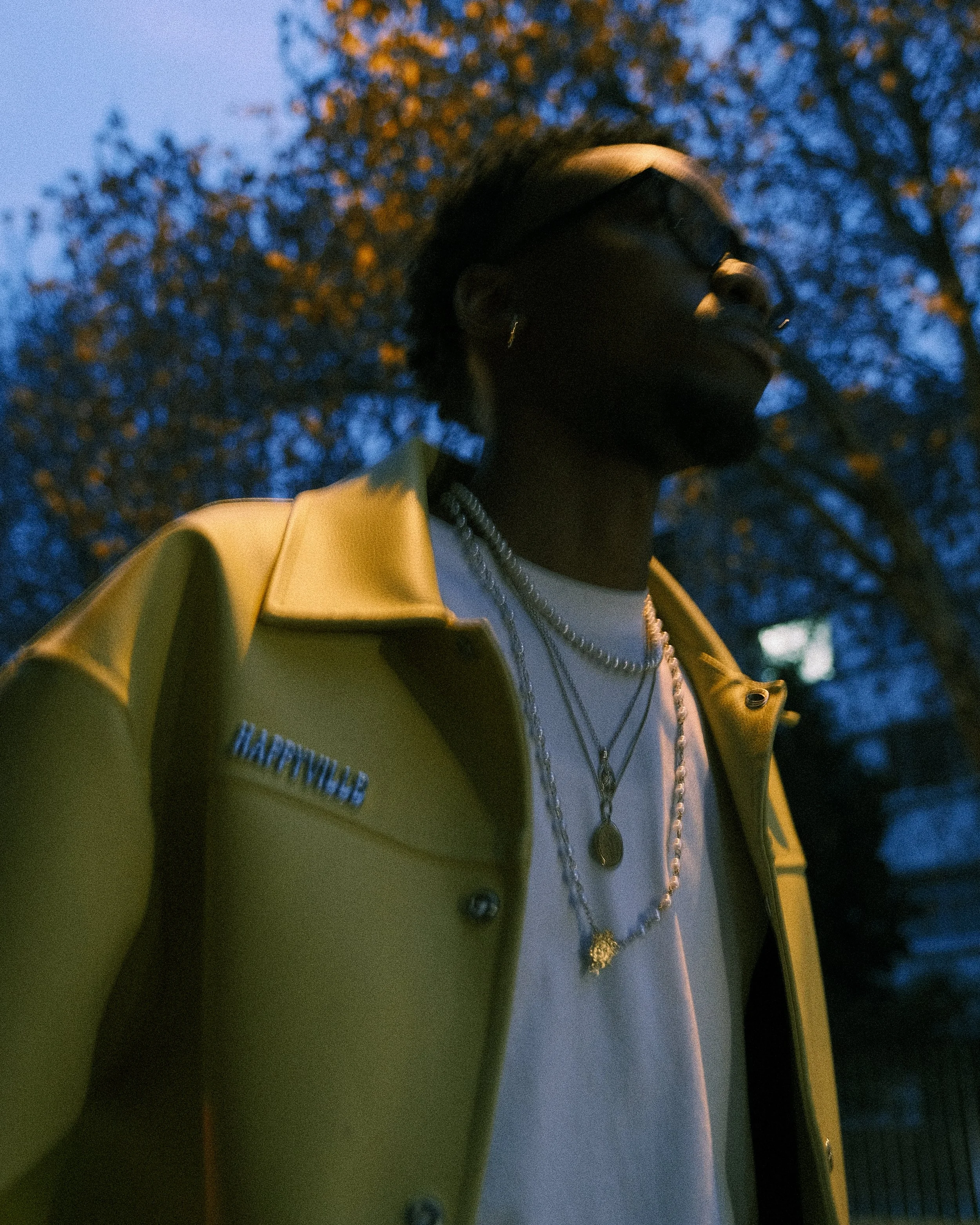 A man wearing a yellow jacket with 'HAPPYFILL' embroidered on the lapel, layered necklaces, a white shirt, glasses, and earrings, standing outdoors at dusk with trees and a darkening sky in the background.