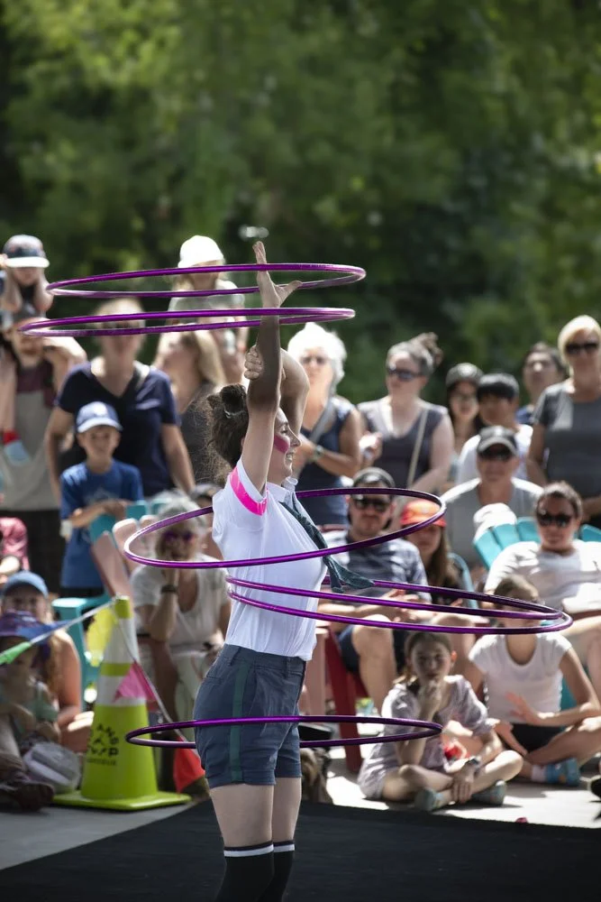 Une jongleuse avec plusieurs cerceaux roses, en train de performer devant une foule d'admirateurs dans un parc en plein air.