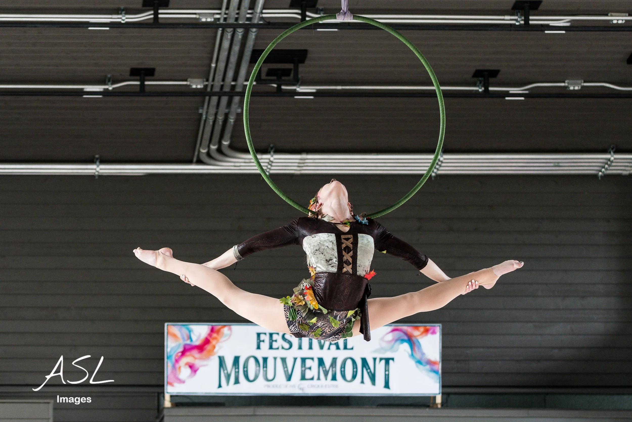 Une artiste de cirque en costume effectue une acrobatie aérienne suspendue à un cerceau vert, en position horizontale, lors d'un festival à Mouvement, avec un panneau en arrière-plan.