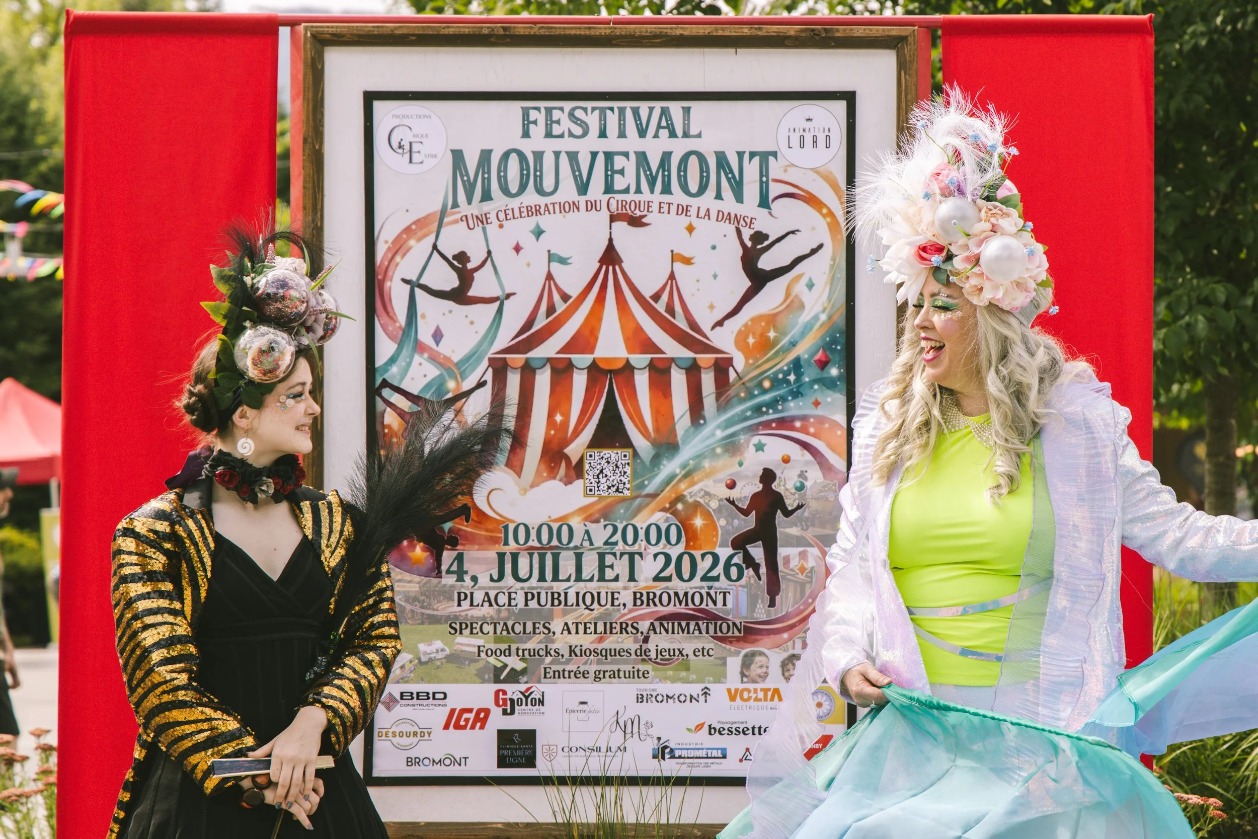 Affiche du festival Mouvement avec deux femmes déguisées en artistes de cirque ou de spectacle de danse, portant des costumes colorés et originales, devant un panneau publicitaire indiquant la date, l'heure et le lieu de l'événement, avec un décor de fêtes en plein air.