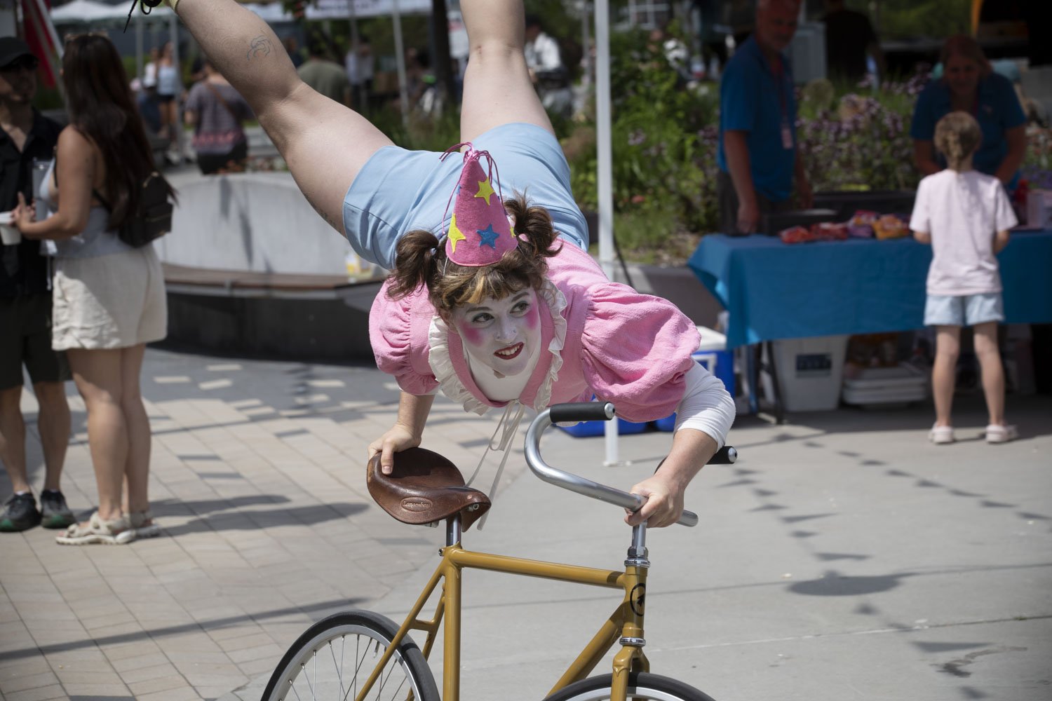 Un artiste clown effectuant une acrobatie sur un vélo lors d'une fête ou événement en plein air, avec des spectateurs en arrière-plan.