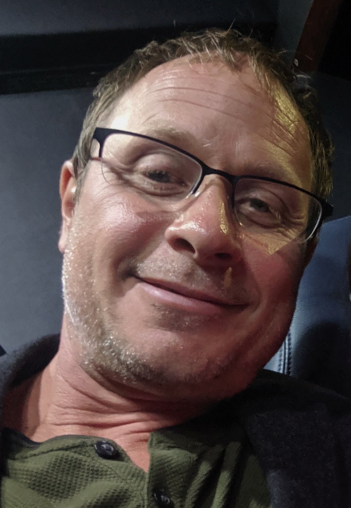 A middle-aged man with glasses, smiling and relaxed. He has short, light brown hair and is wearing a dark shirt with a collar.
