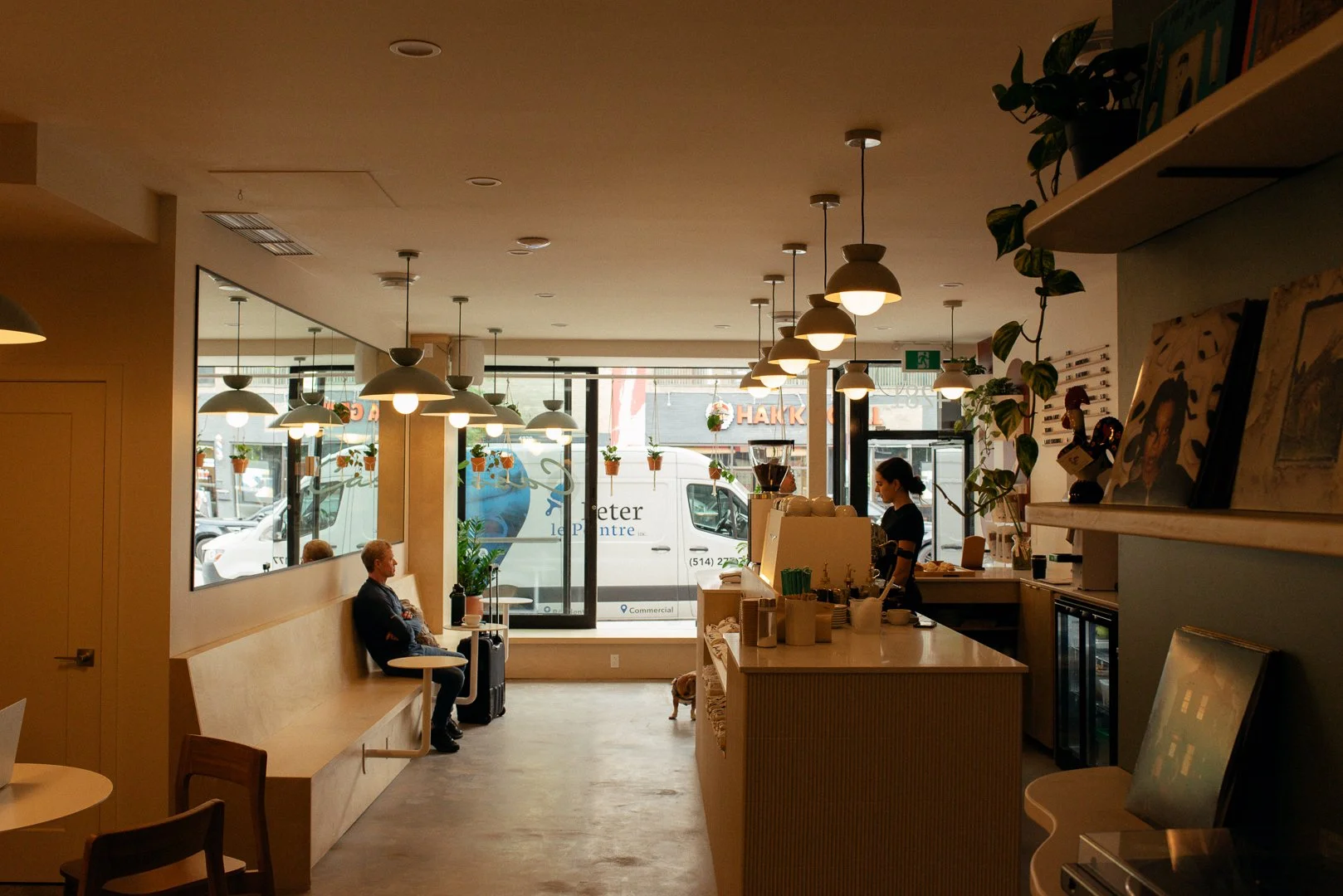Interior of a cozy coffee shop with hanging pendant lights, a counter with a barista, and large front windows showing parked vehicles outside.
