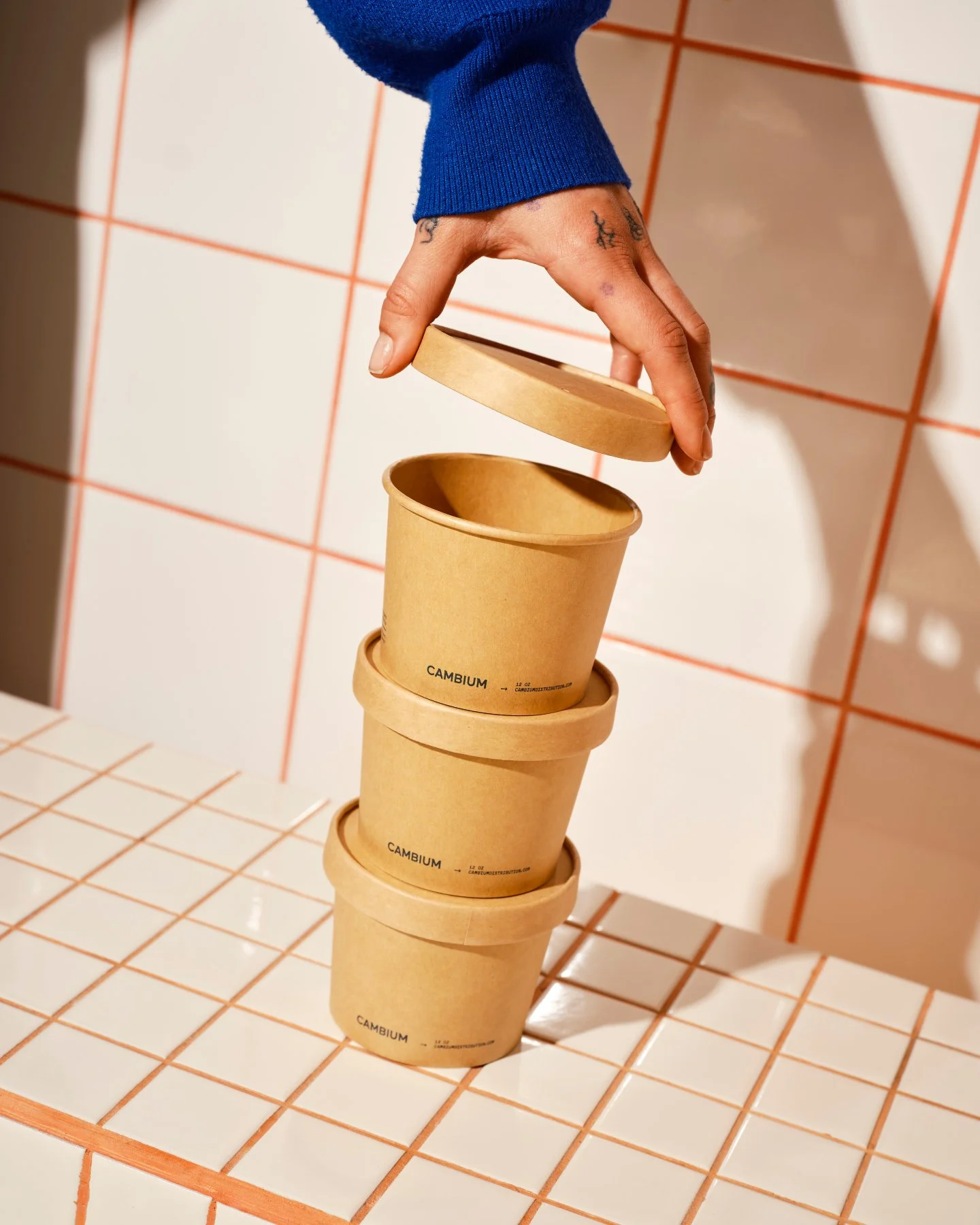 A hand with tattoos is lifting the lid off a stack of three cardboard containers. The containers are stacked on a tiled surface with orange grout lines, and a tiled wall with the same color grout lines is in the background.