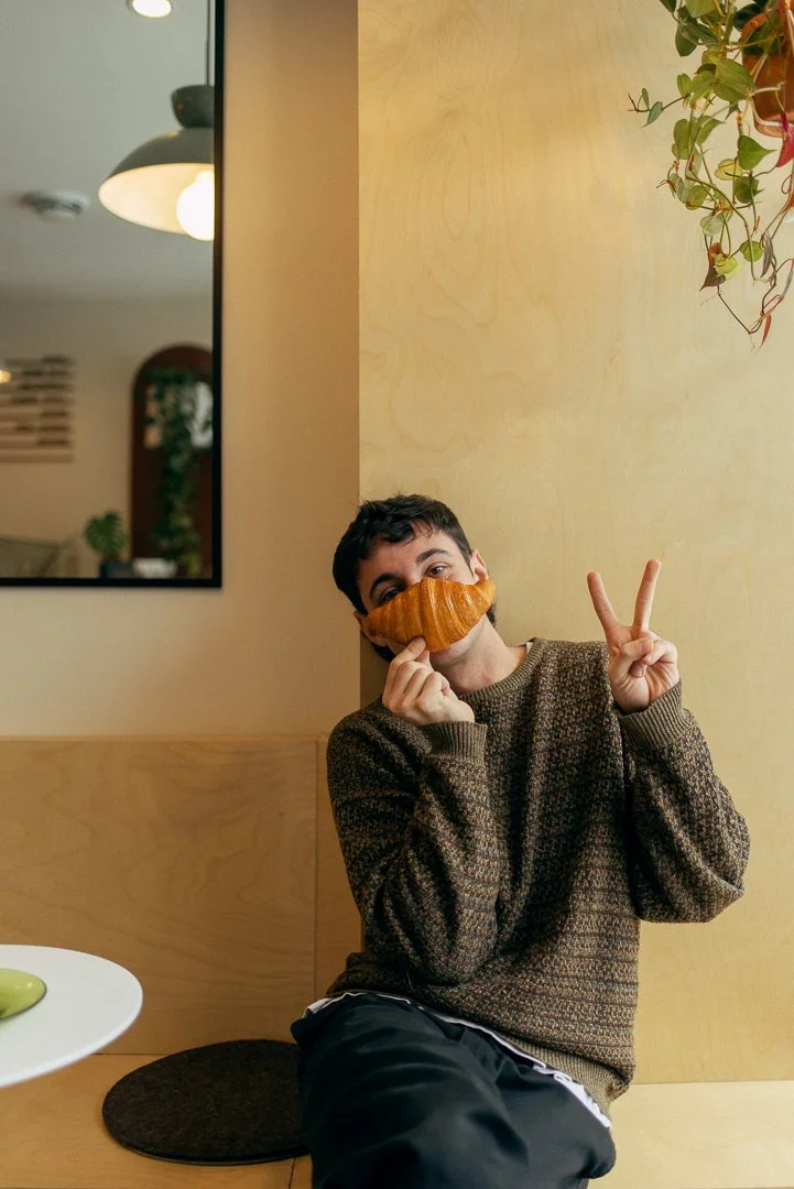 Young man in a grey polo shirt and cream-colored pants sitting on a beige cushioned bench with a beige wall background.
