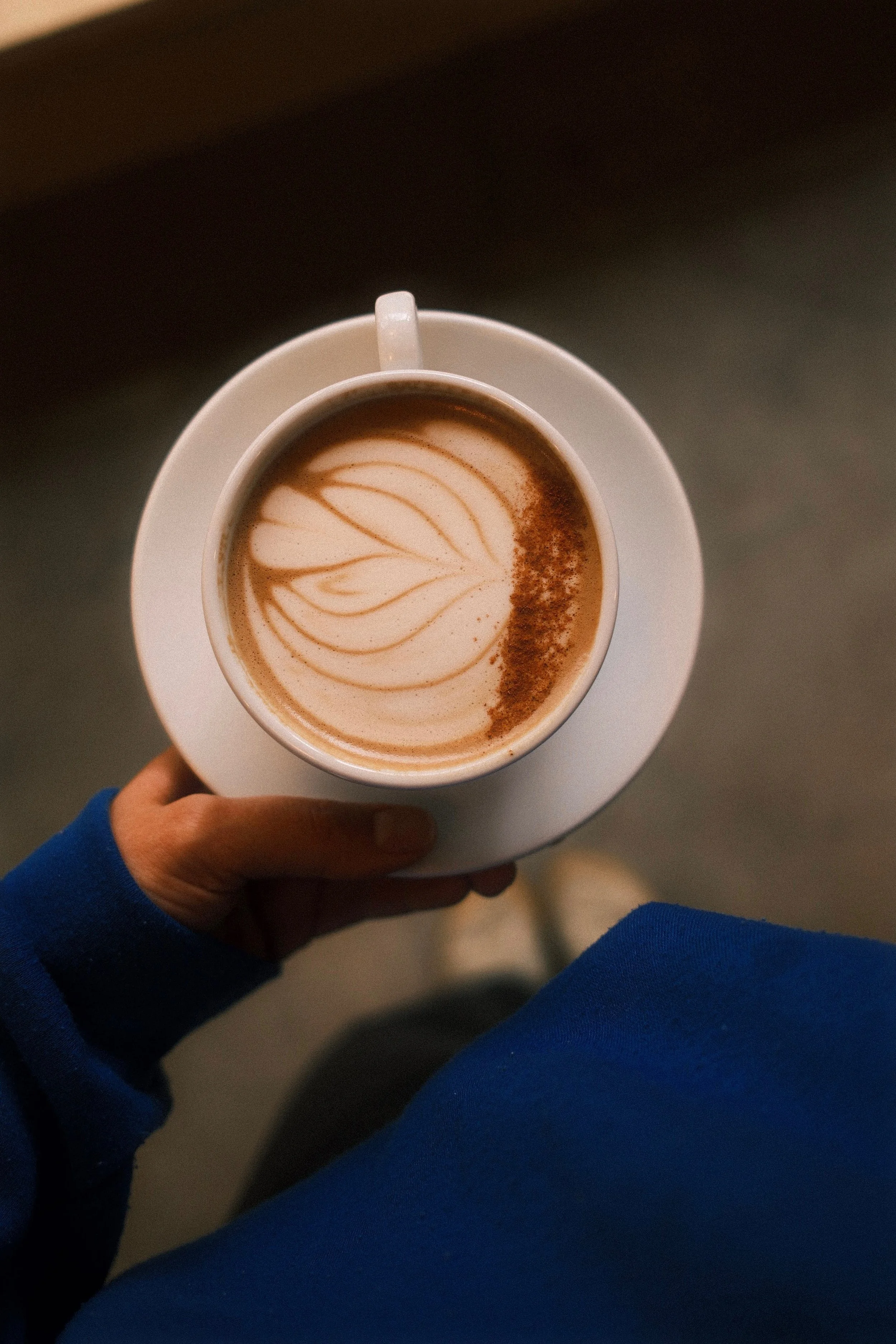 Top-down view of a person holding a white cup of latte with latte art on top, sitting on a white saucer on a wooden surface.