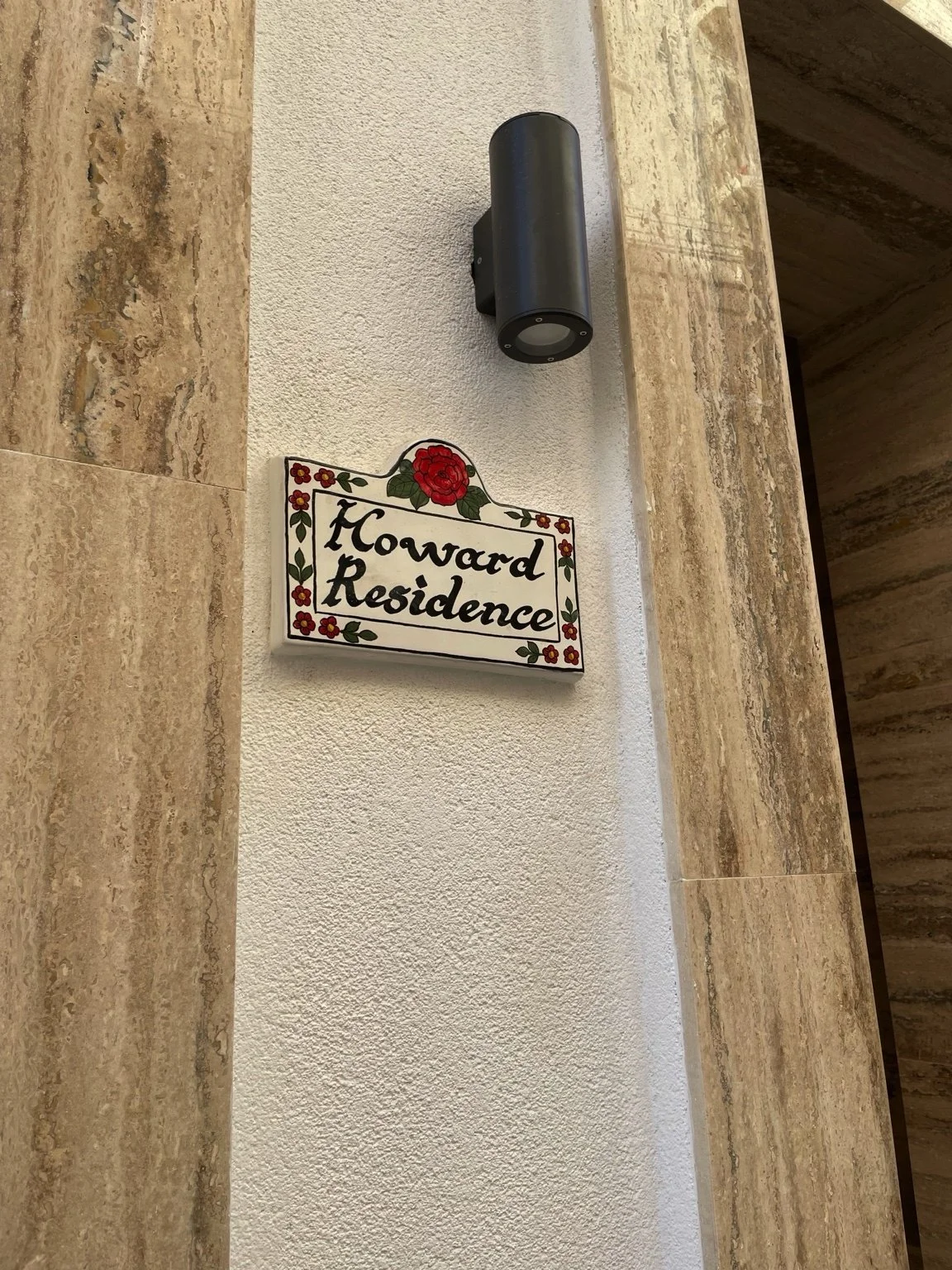 A decorative wall sign that reads 'Howard Residence' with a red rose and small red flowers, mounted on a wall next to a modern outdoor wall light fixture.
