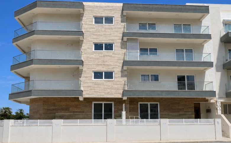 A modern apartment building with four floors, glass balconies, and a mix of white and beige exterior walls, set against a clear blue sky.