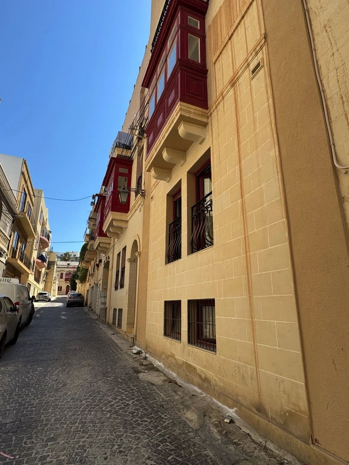 A narrow street lined with beige and cream-colored buildings with balconies, some with black wrought iron railings, under a clear blue sky. Cars are parked along the street and laundry is hanging from balconies.