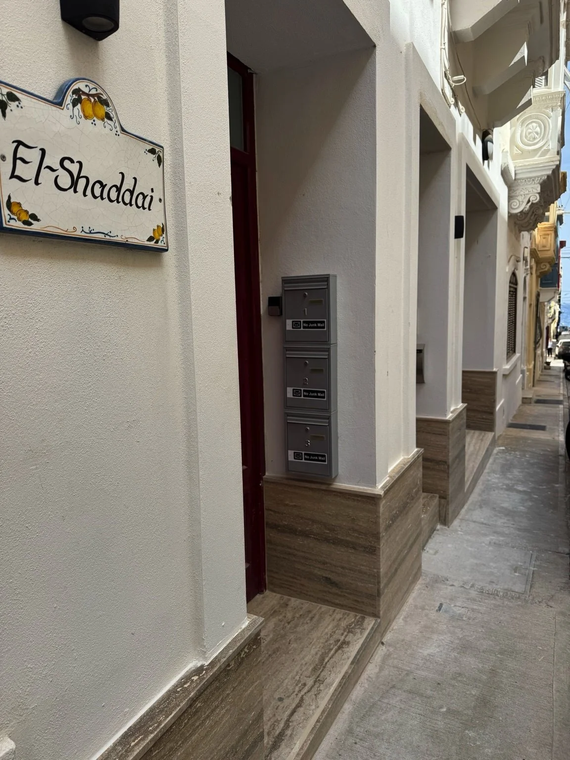 Outdoor sidewalk with white building wall, a sign with yellow lemon illustrations reading 'El-Shaddai', and three gray mailboxes mounted on the wall.