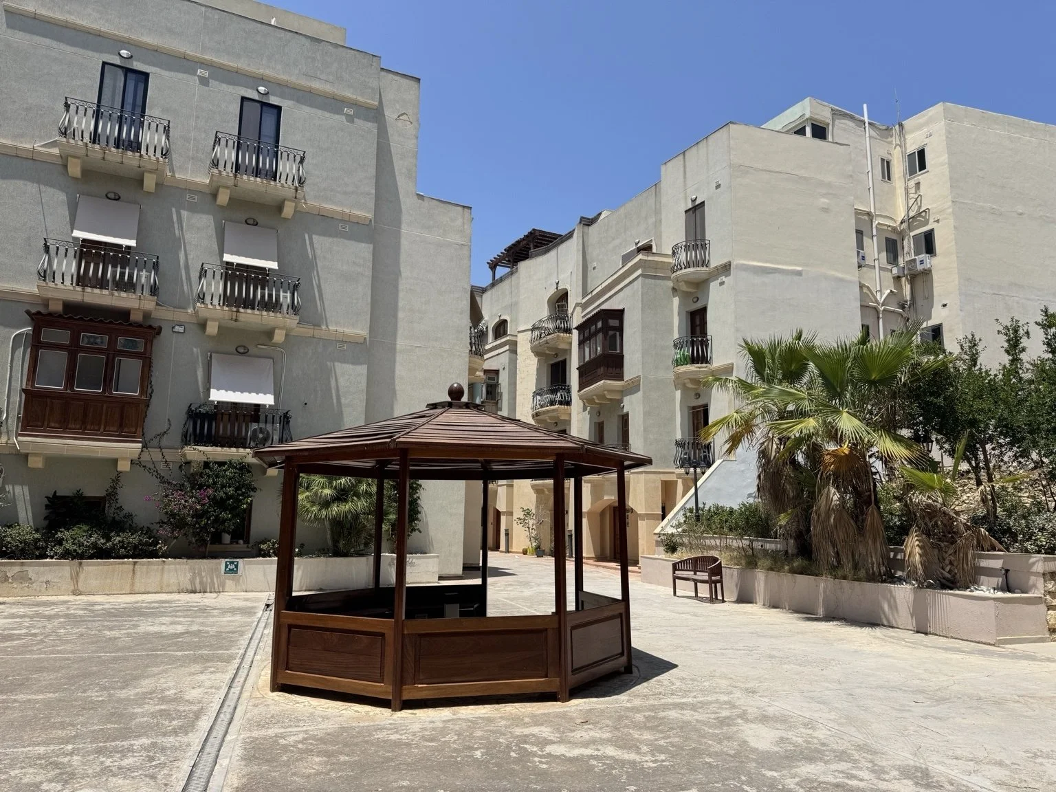 An apartment courtyard with a wooden gazebo, benches, palm trees, and white multi-story buildings with balconies under a blue sky.