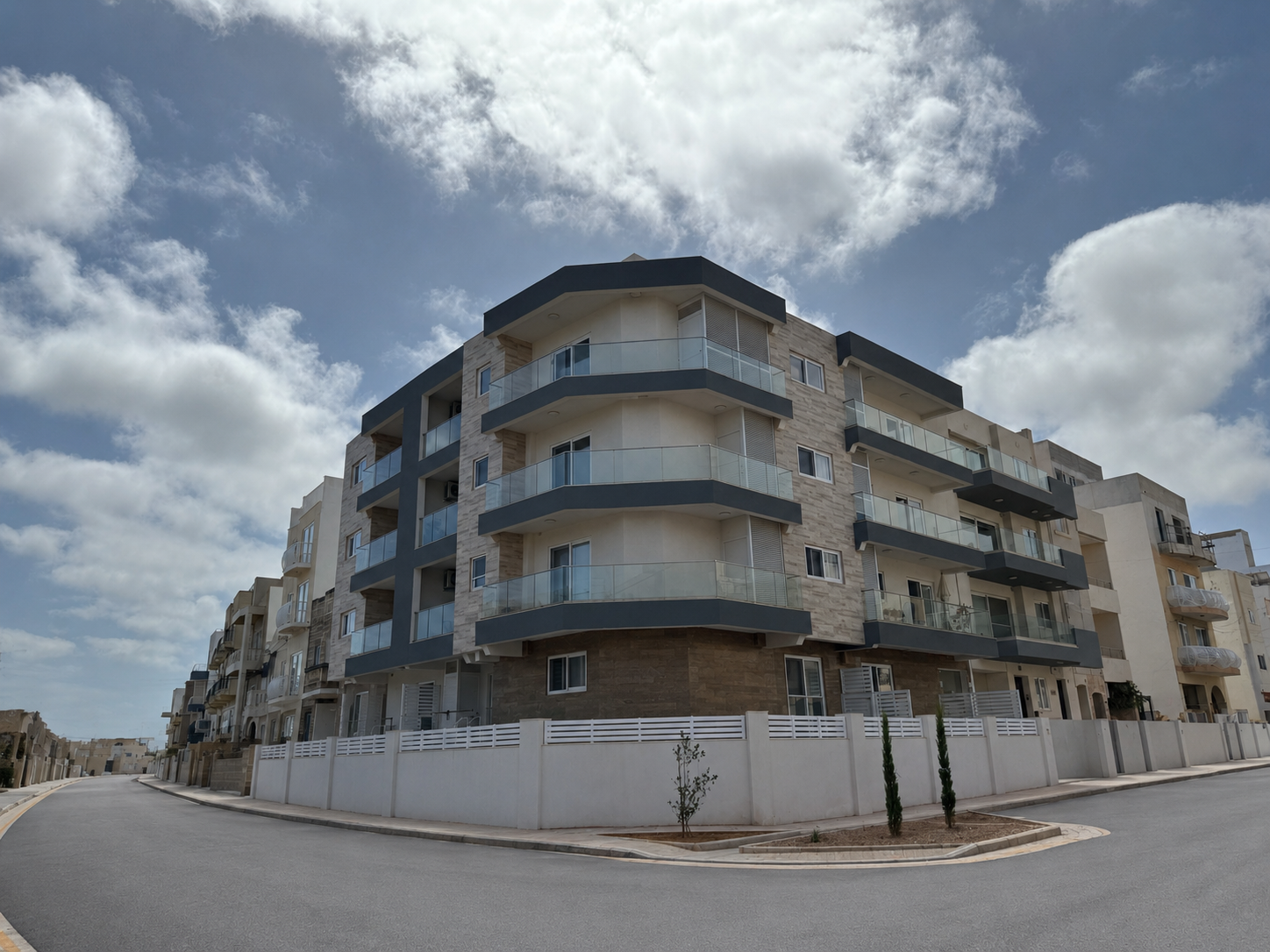 A modern four-story residential building with balconies and glass railings, situated on a curved street under a partly cloudy sky.
