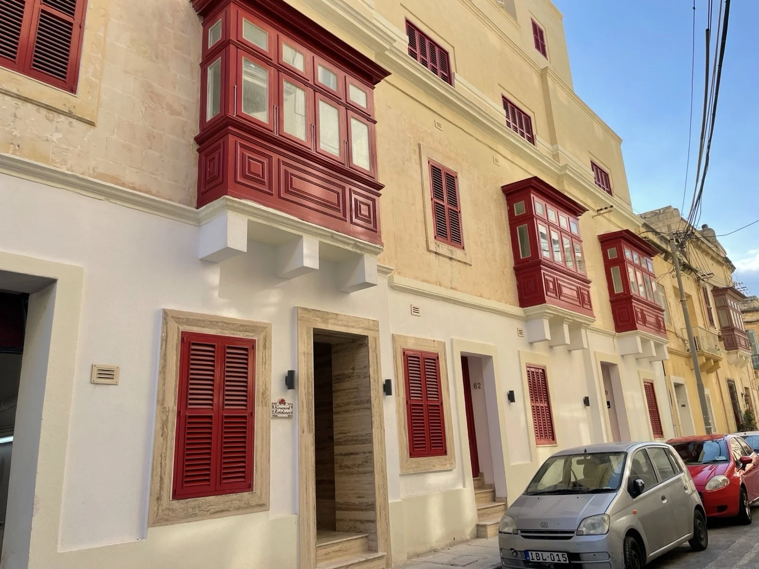 A row of multi-story buildings with beige and cream-colored facades, red window shutters, and red balconies on the second floor. The street has parked cars, including a silver hatchback in the foreground. Power lines run along the street, and the sky is clear and blue.
