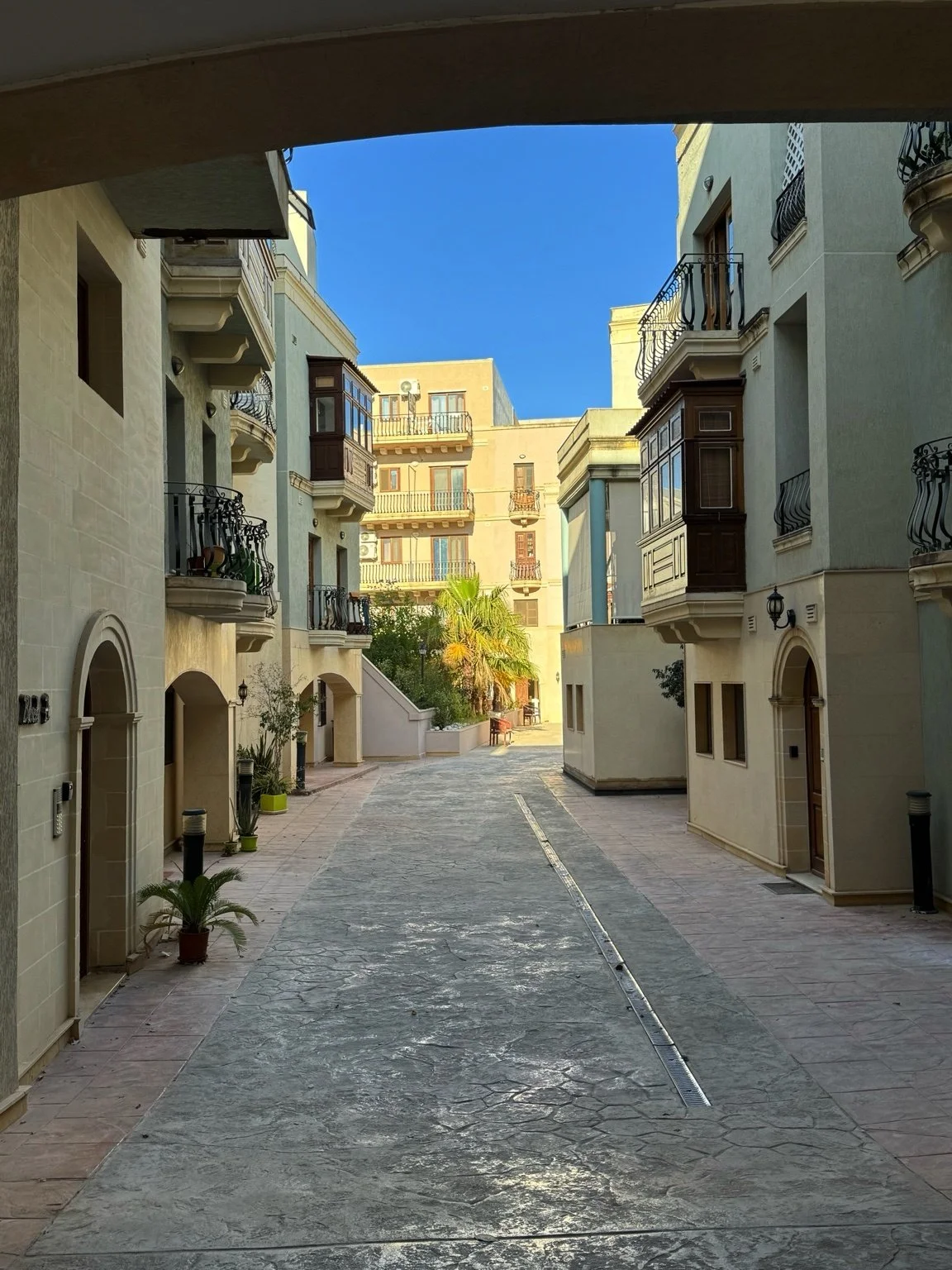 A quiet courtyard surrounded by beige and cream-colored multi-story buildings with balconies and black wrought-iron railings, some with plants. Clear blue sky overhead, with a small palm tree and other greenery. The ground has a stone pathway with a metal drainage channel.