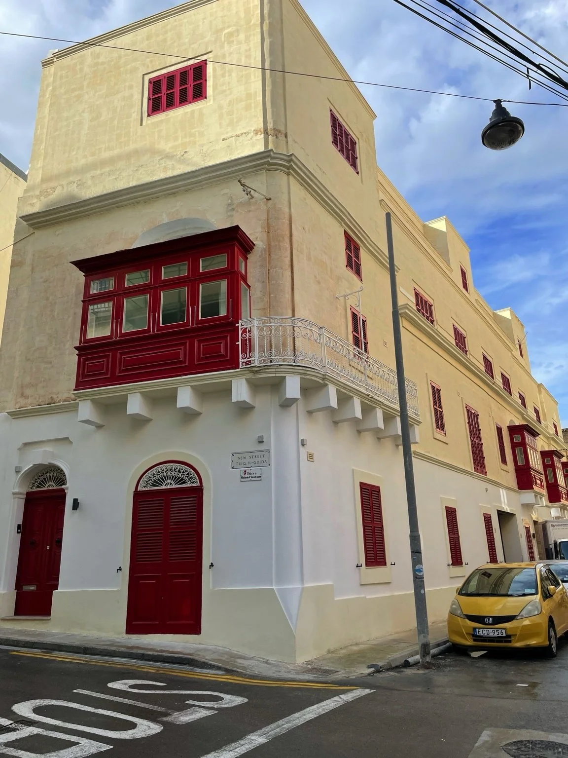 Street view of a Mediterranean-style building with cream-colored walls, red shutters, and red windows. A corner balcony with a white decorative railing and a red enclosed porch are visible. A street sign reads 'New Street Tieg Il-Gudda.'