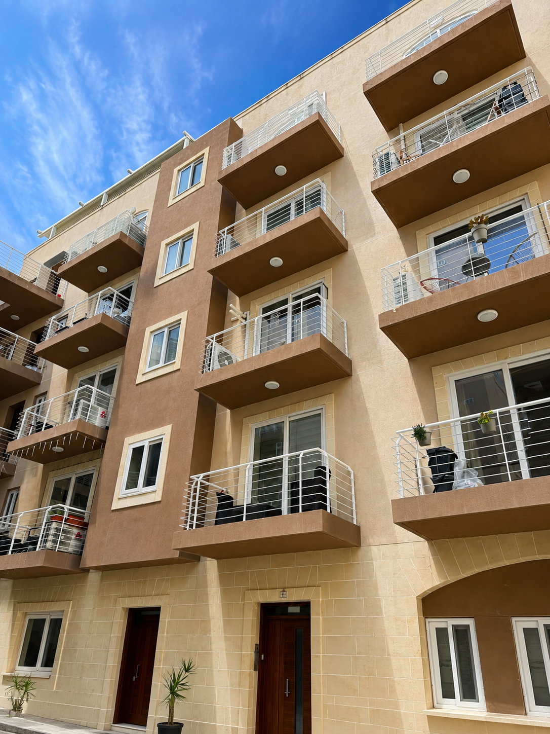 Exterior view of a modern apartment building with multiple balconies, some having potted plants and furniture, against a blue sky with clouds.