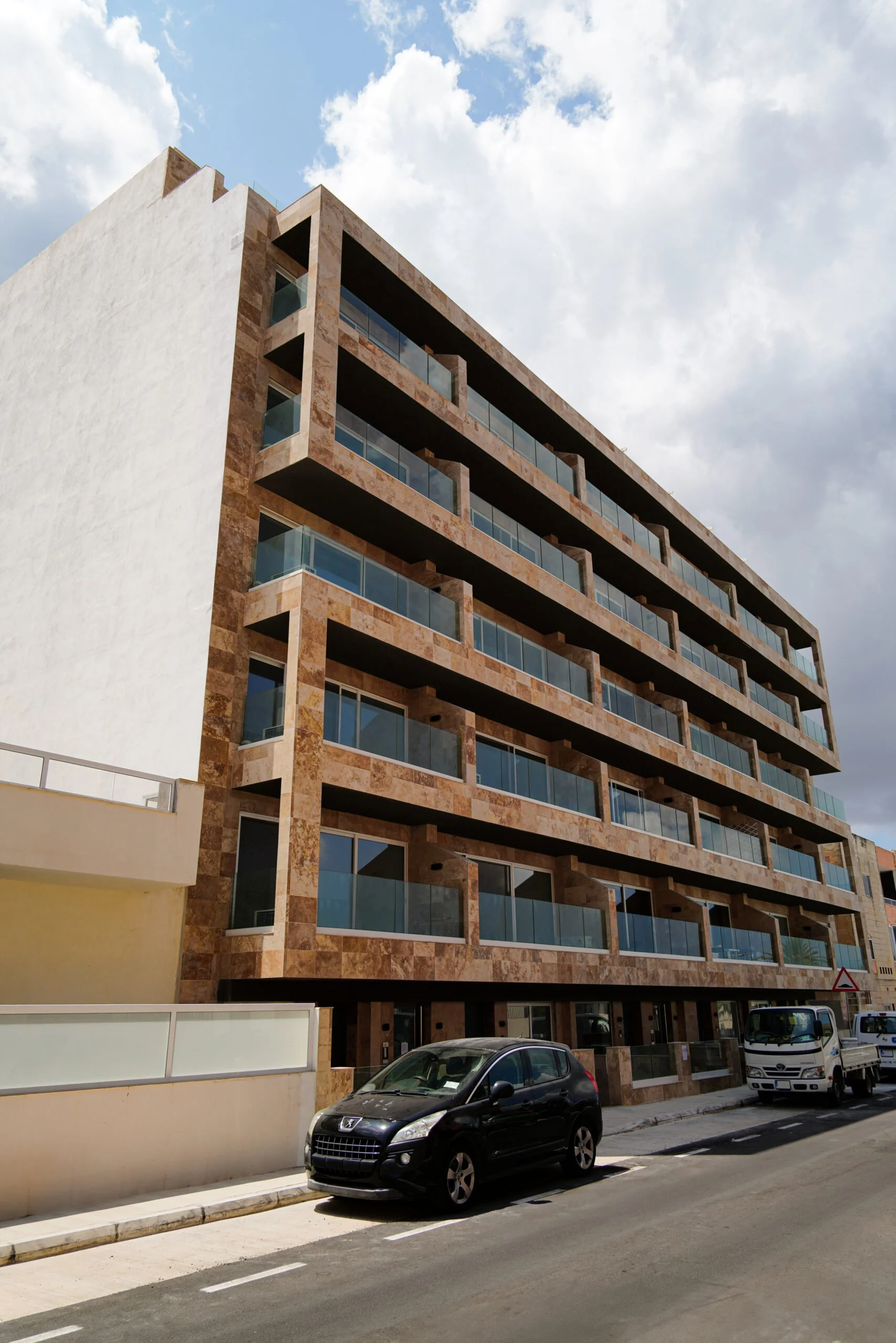 Modern multi-story apartment building with balconies and glass railings, parked cars in front, under a partly cloudy sky.