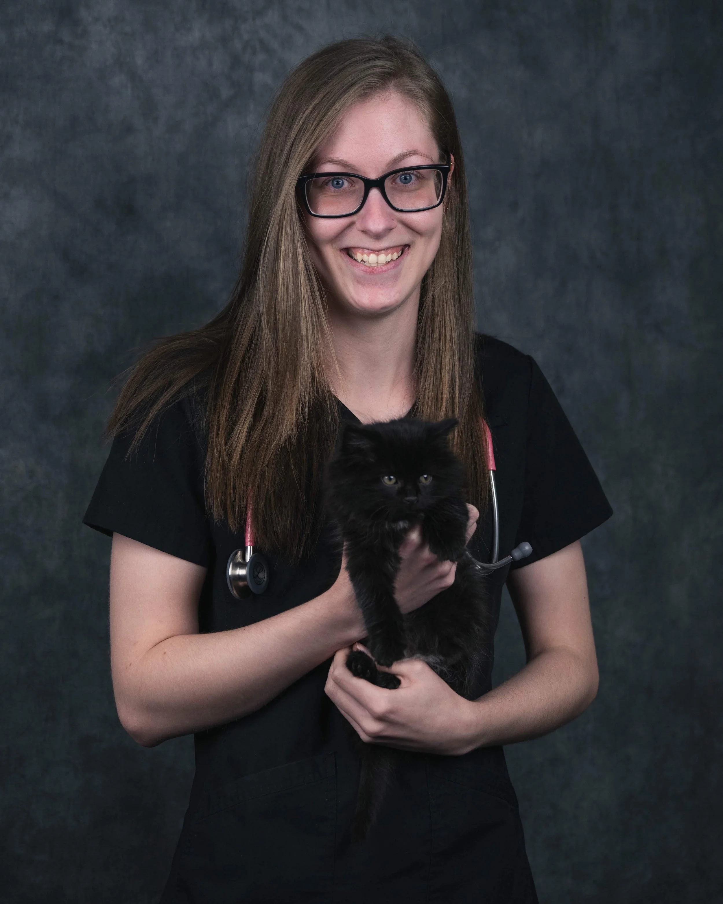 Jeune femme souriante qui prend la pose avec un petit chat noir dans les mains. Elle est en tenu de scrub avec u stéthoscope autour du cou. C'est ça photo de graduation pour sa technique en santé animale