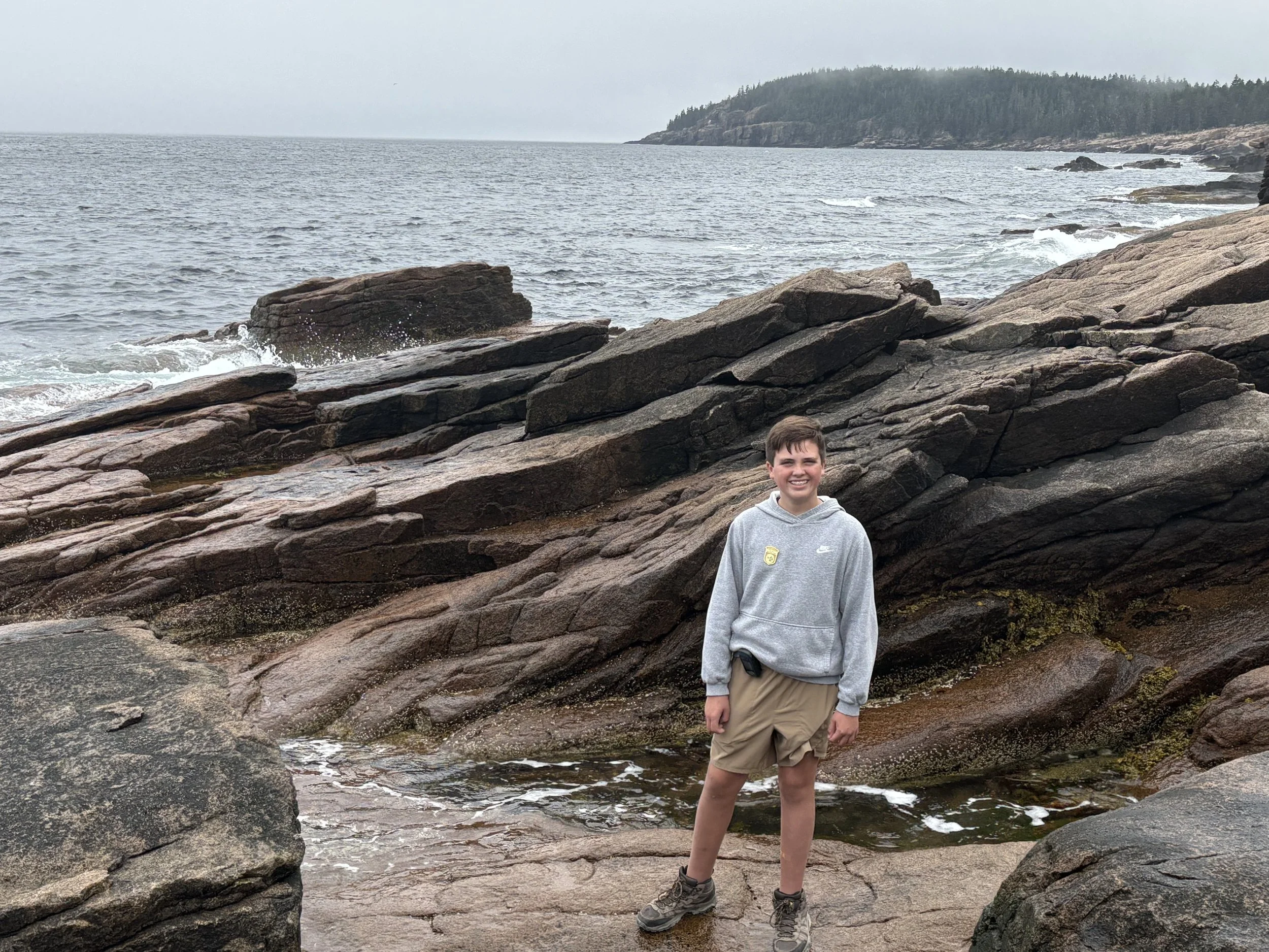 A young boy stands on a rocky shoreline with the ocean in the background. He is wearing a gray hoodie, khaki shorts, and hiking boots, smiling at the camera.