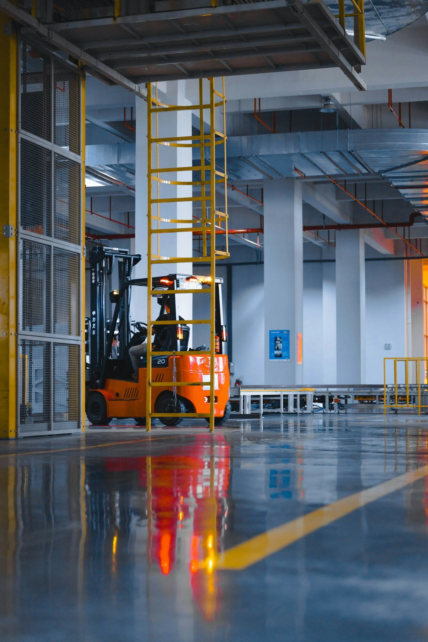 Ein orangener Gabelstapler fährt durch eine Lagerhalle mit metallicfärbigen Lüftungsschächten an der Decke und gelben Sicherheitsgeländern. Die Halle ist leer und gut beleuchtet, das reflektierende Boden zeigt die Lichter und Strukturen.
