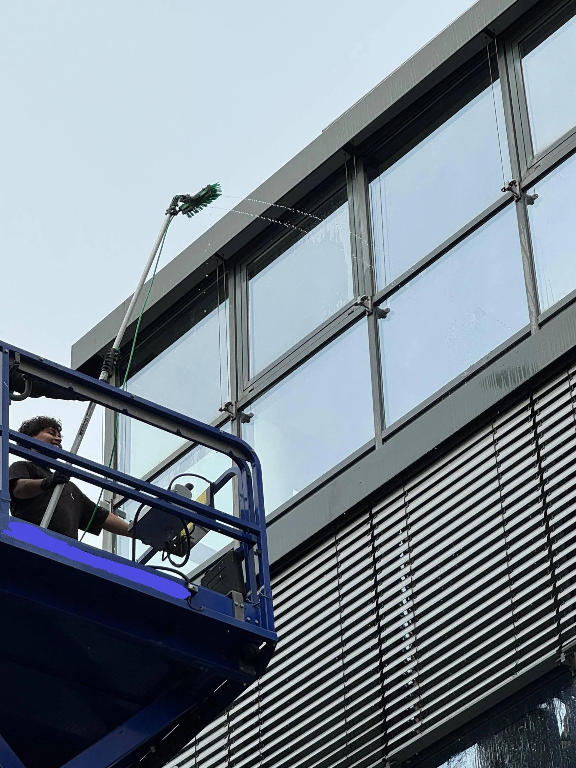 Ein Mann steht auf einem Hochhubwagen und reinigt die Fenster eines modernen Gebäudes mit einer Scheuerbürste.