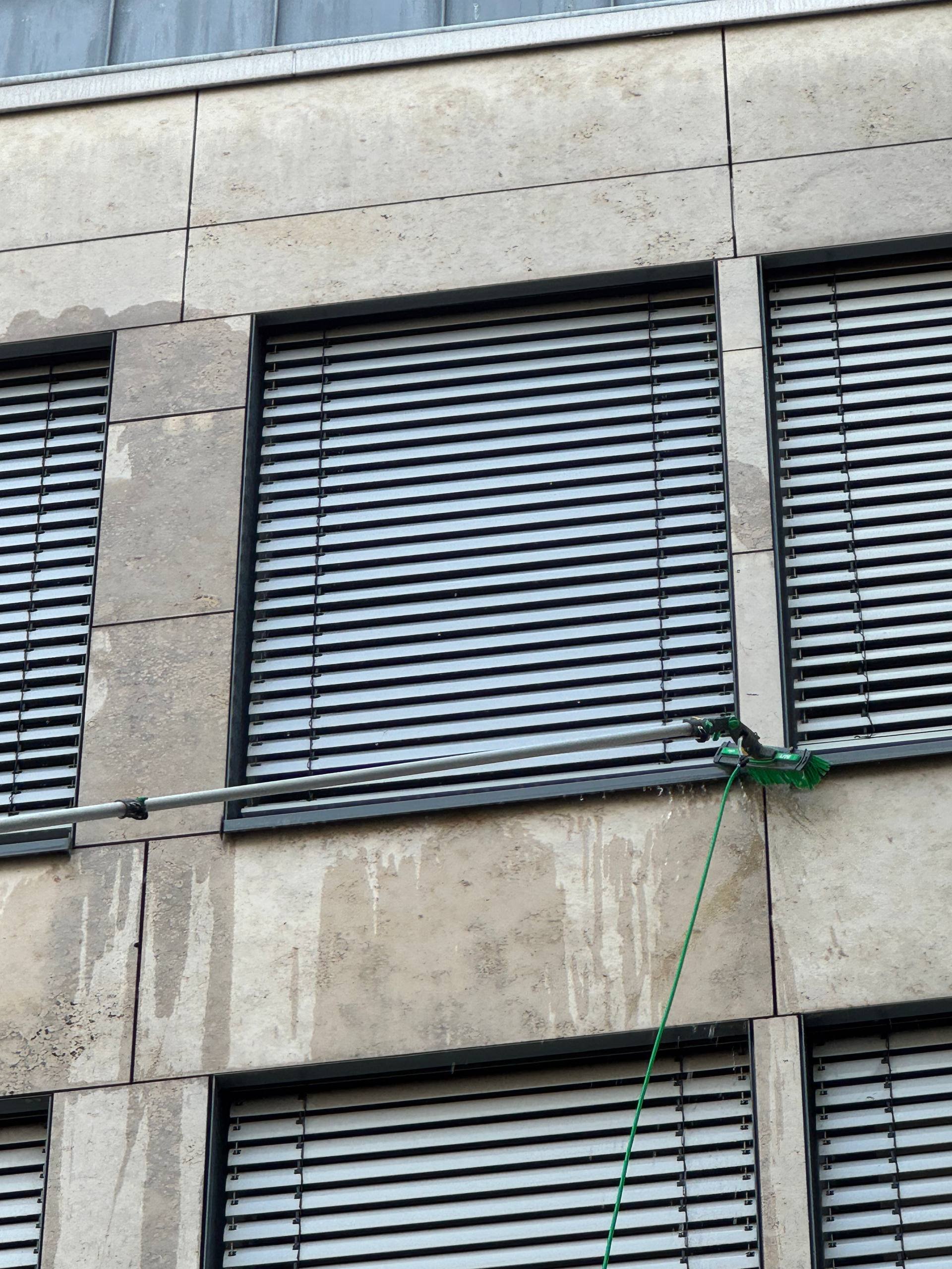 Die Fassade eines modernen Gebäudes mit behinderten- und umweltfreundlichen Elementen, darunter Fenster mit Jalousien und eine Reinigungsbürste an einem langen Stiel, die an der Wand befestigt ist.