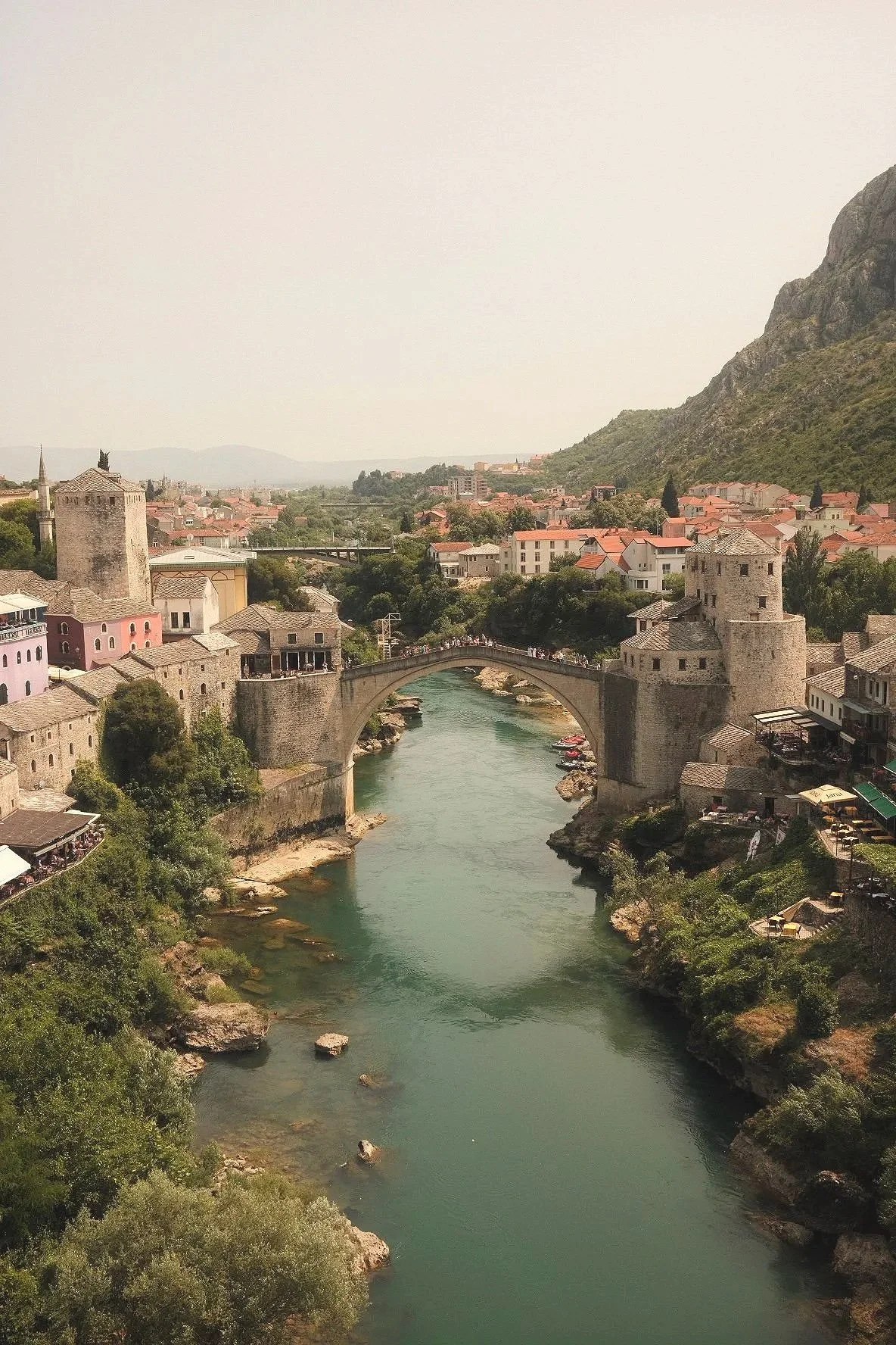 A scenic view of a historic stone bridge crossing a river with old European-style buildings on either side and a mountainous landscape in the background.