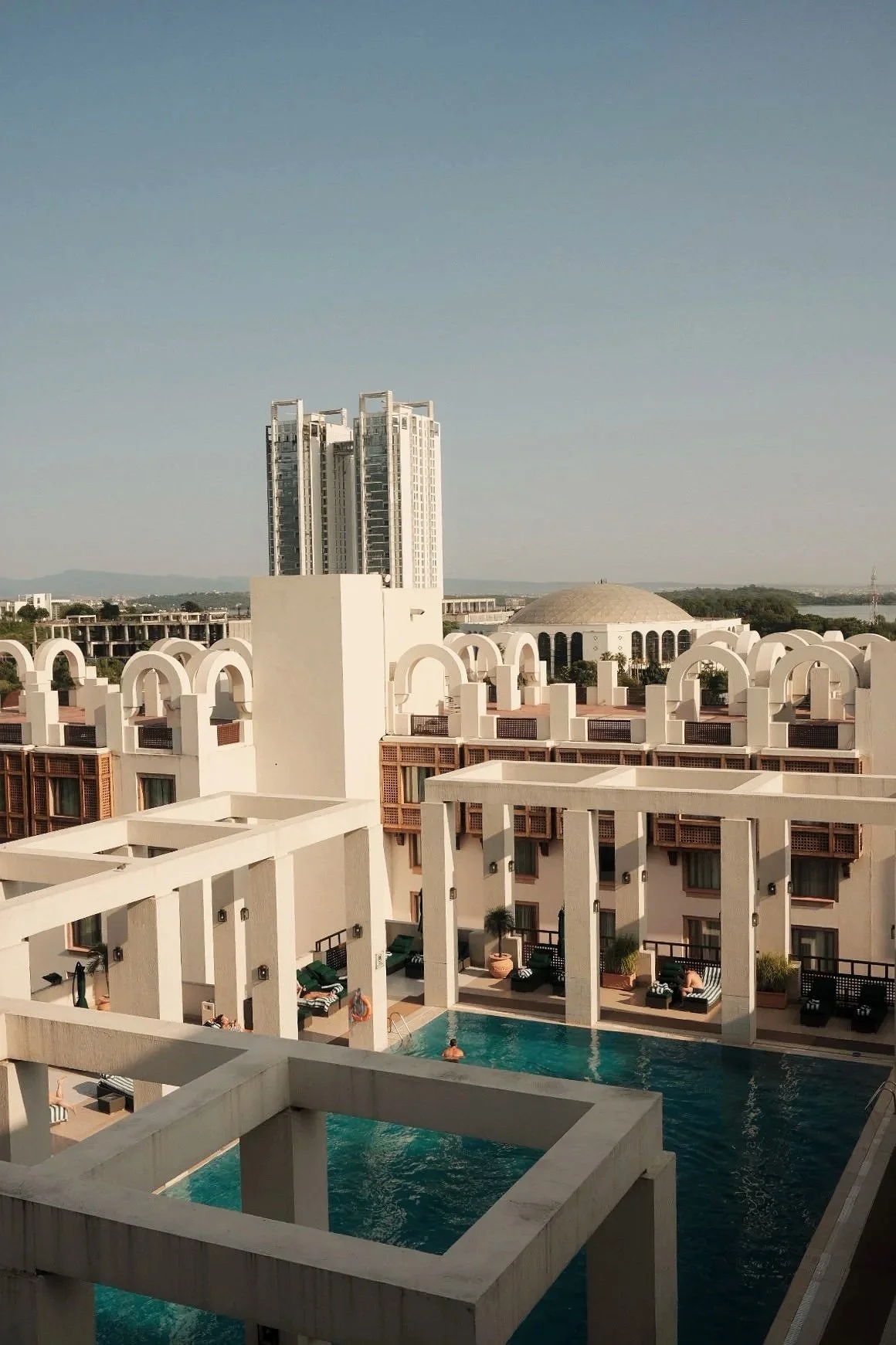 A view of a hotel courtyard with swimming pools, white buildings with arches, and lounge chairs, with a city skyline and a domed building in the background under a clear sky.
