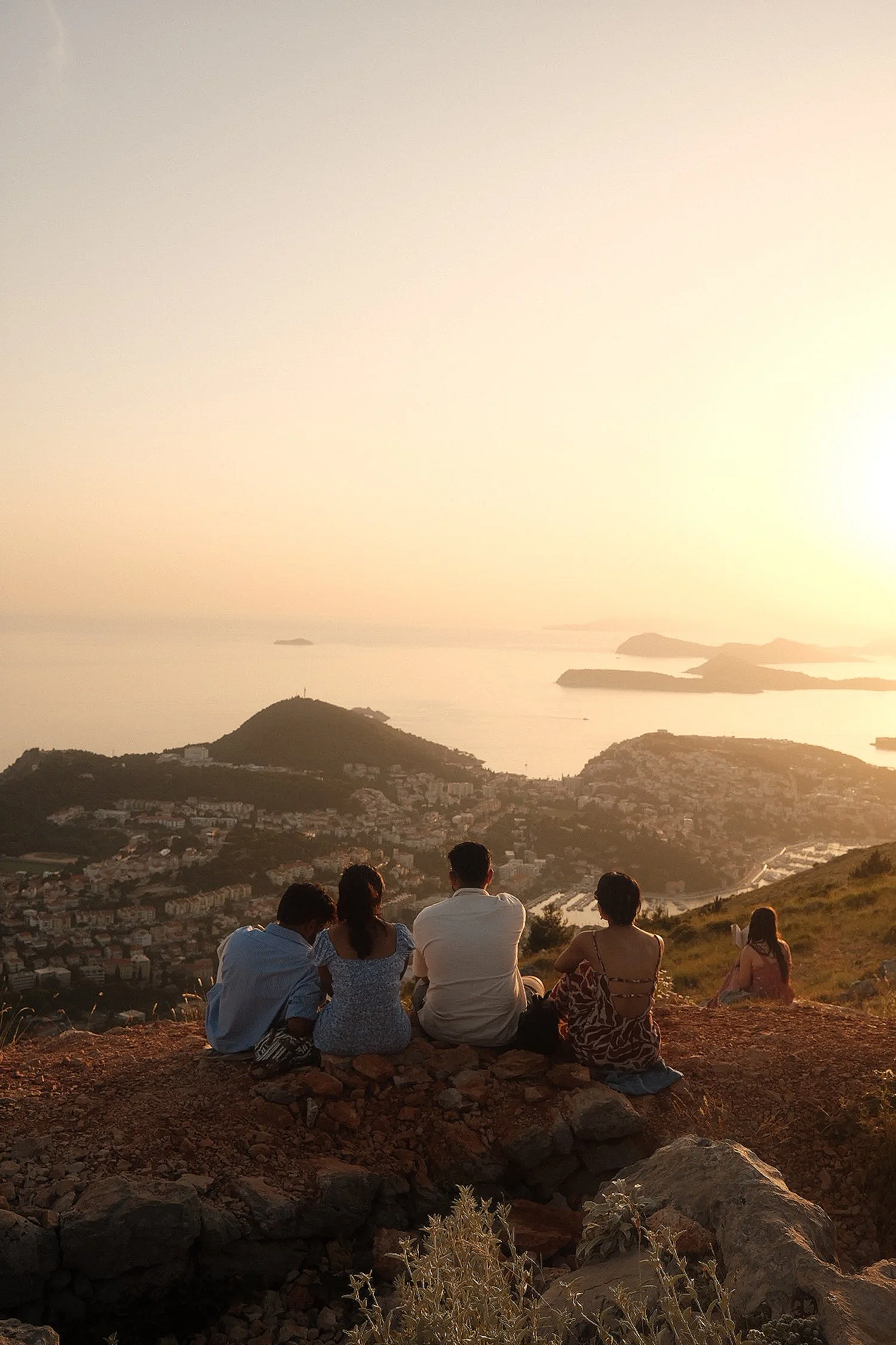 Five people sitting on a hilltop overlooking a city and water at sunset, with the sun low on the horizon and a clear sky.