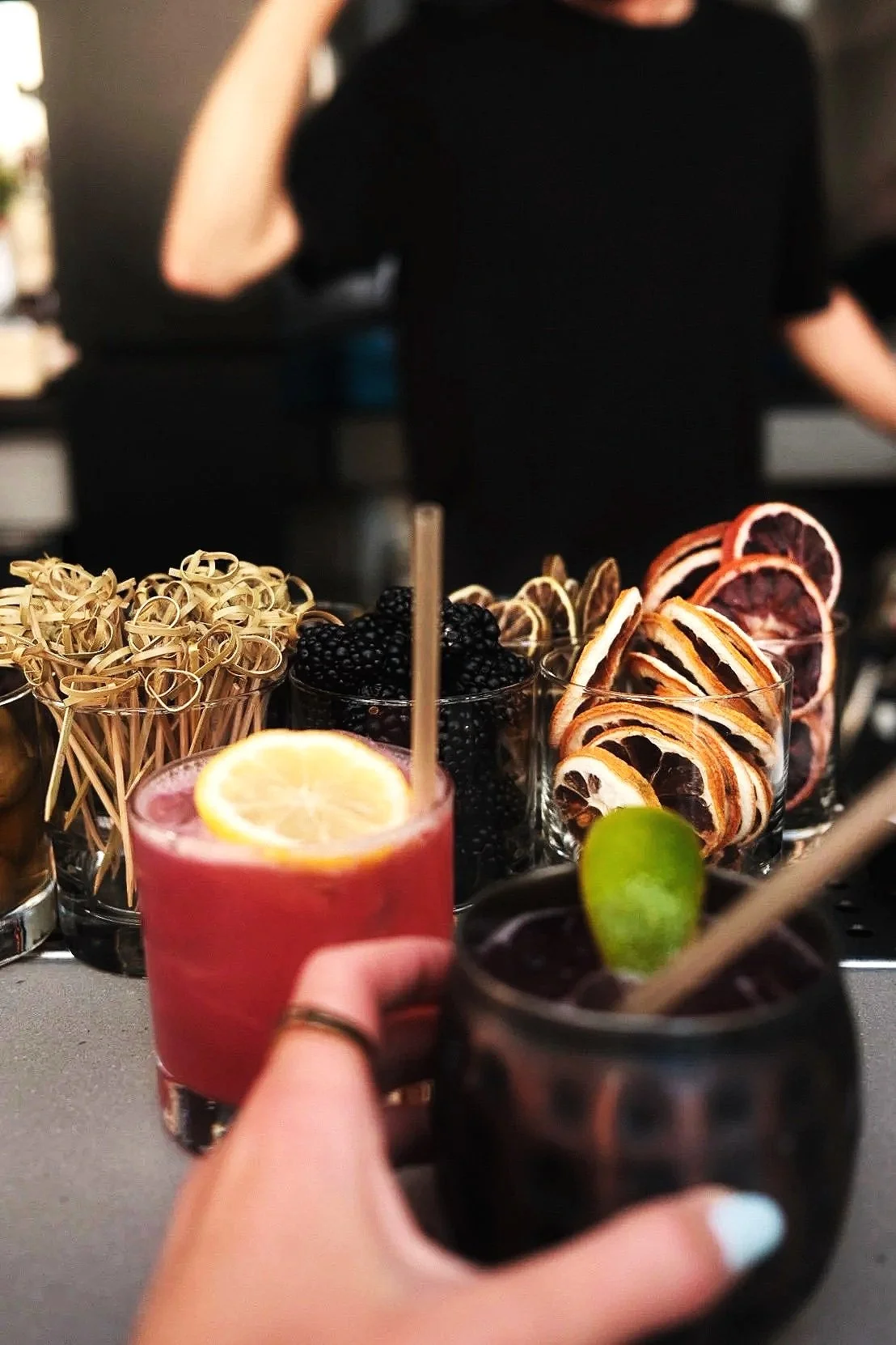 Two cocktails with garnishes in front of a variety of glass desserts and snacks, including blackberries, citrus slices, and patterned cookies, on a bar counter.