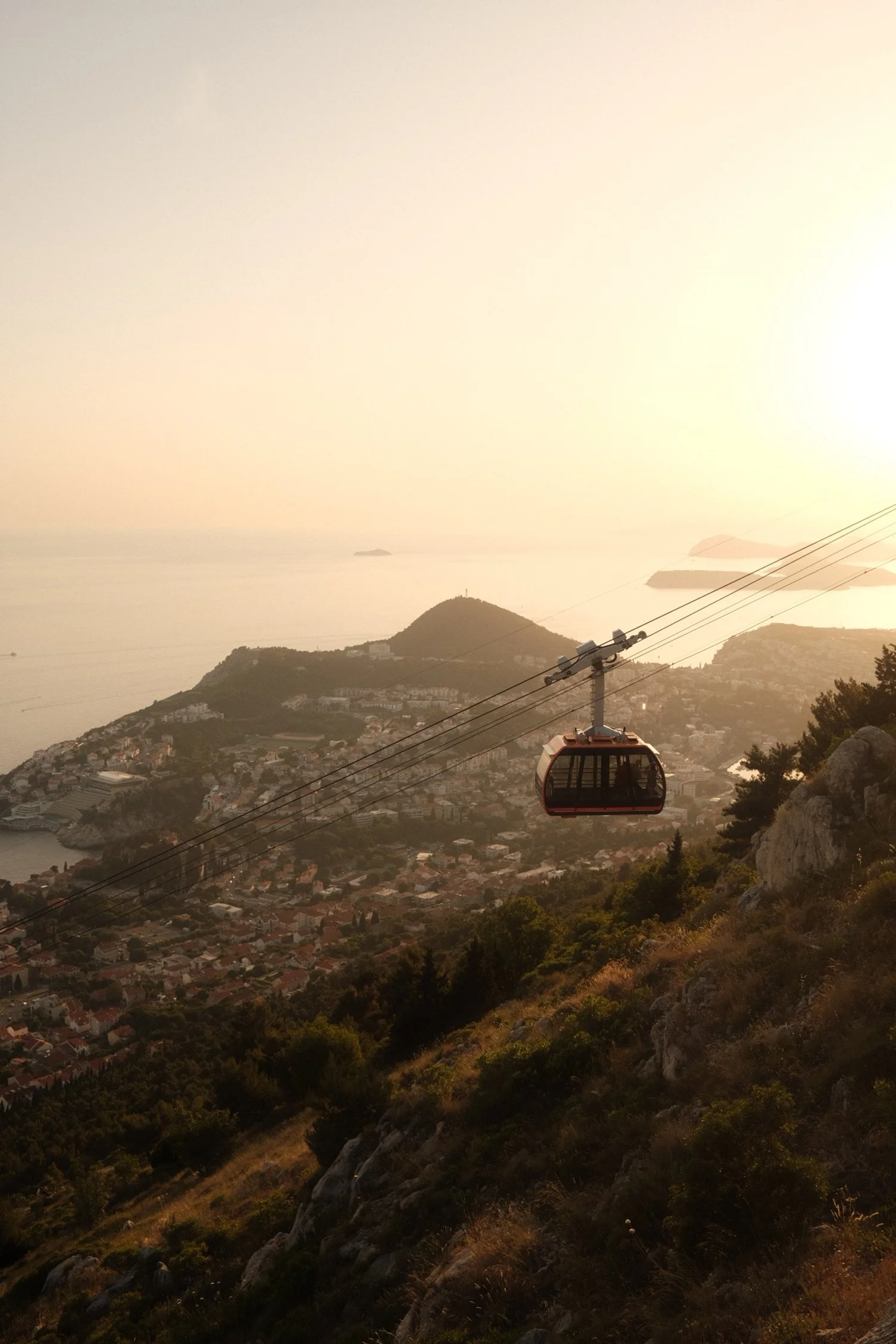 Cable car ascending above a coastal city with hills and water in the background at sunset.