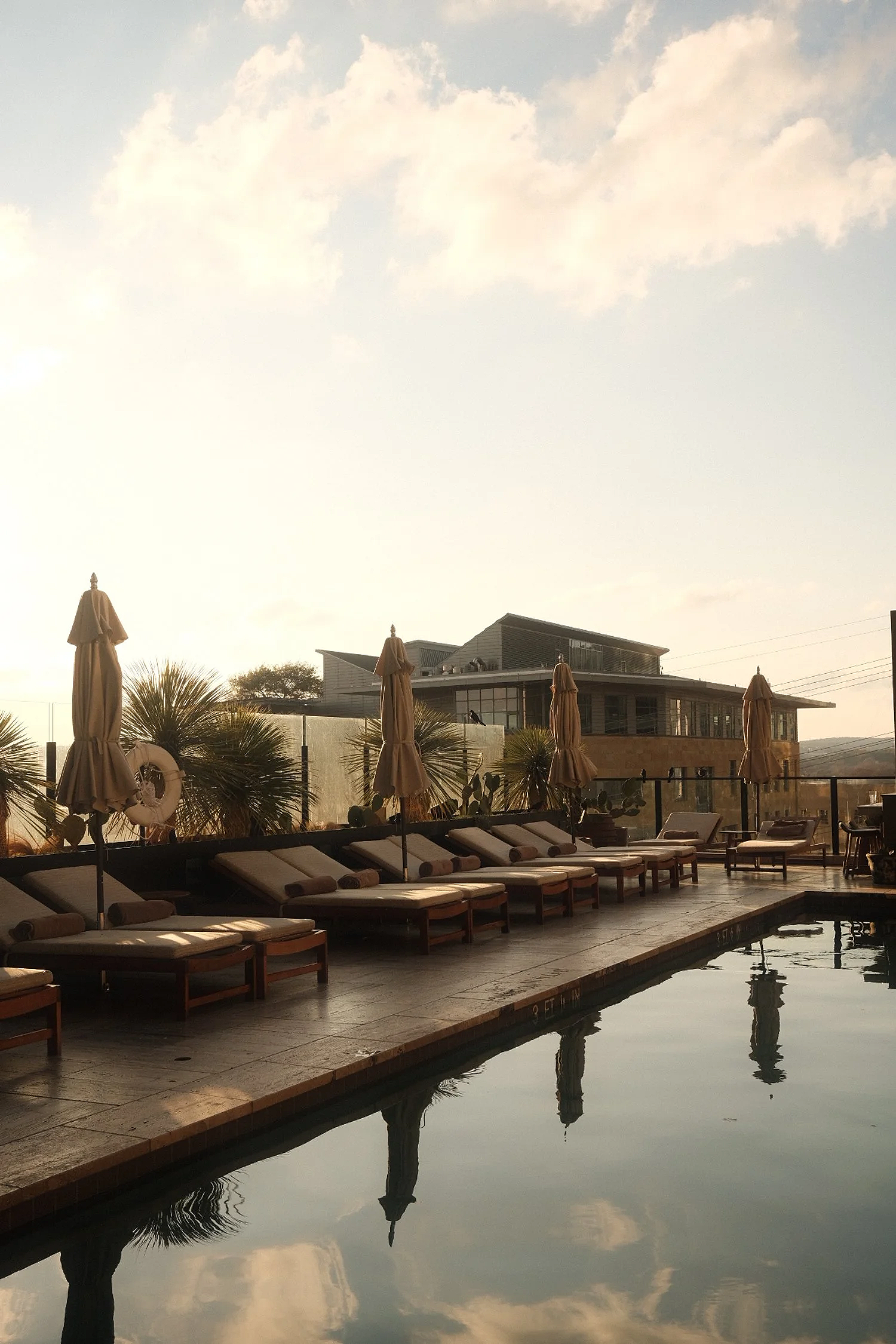 Poolside area with lounge chairs and umbrellas at a resort, with a modern building in the background and cloudy sky overhead.