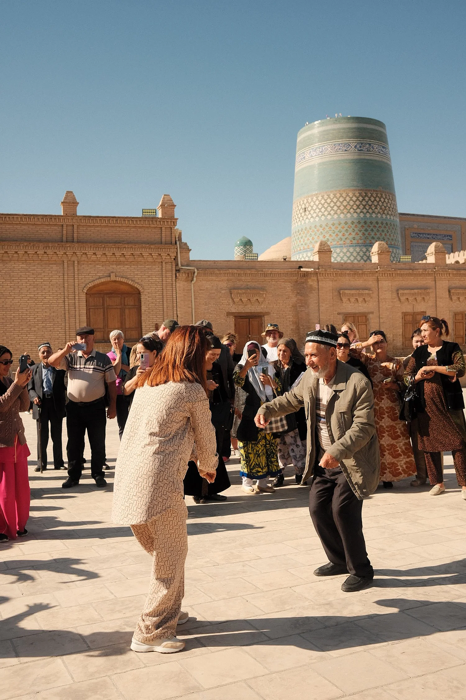 A group of people dancing and taking photos outside a historic building with a mosque dome in the background.